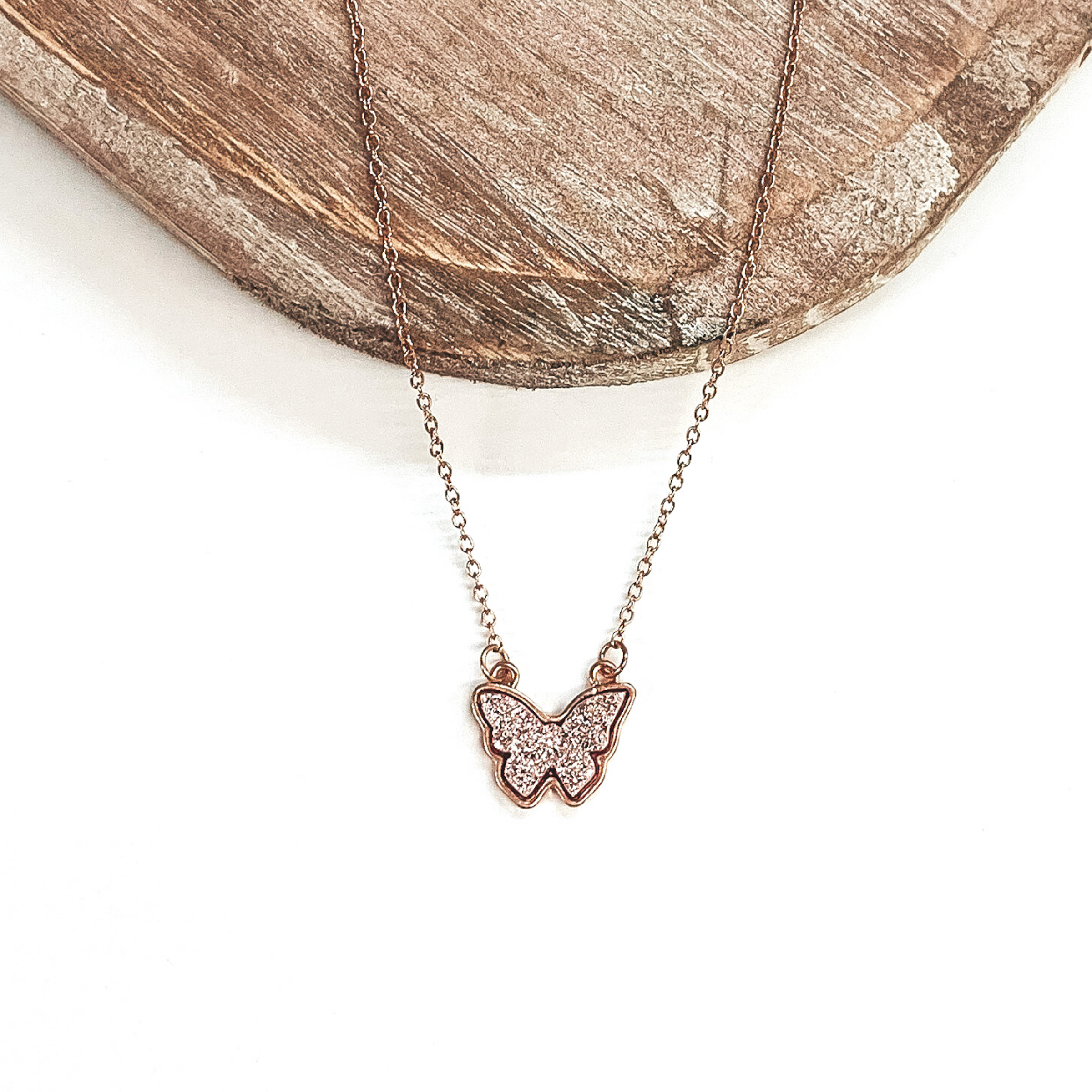 Gold necklace with rose gold druzy butterfly pendant. Pictured on a white and brown background.