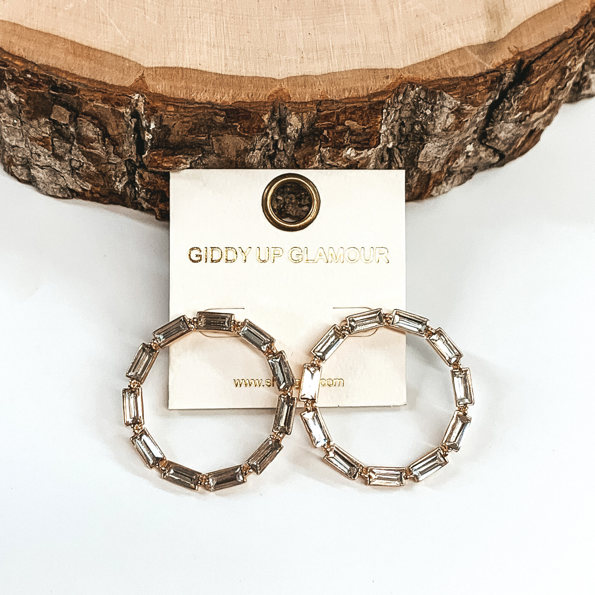 These are gold, open circle studs with emerald shaped clear crystals. These earrings are pictured on a white background with a piece of wood at the top of the picture. 