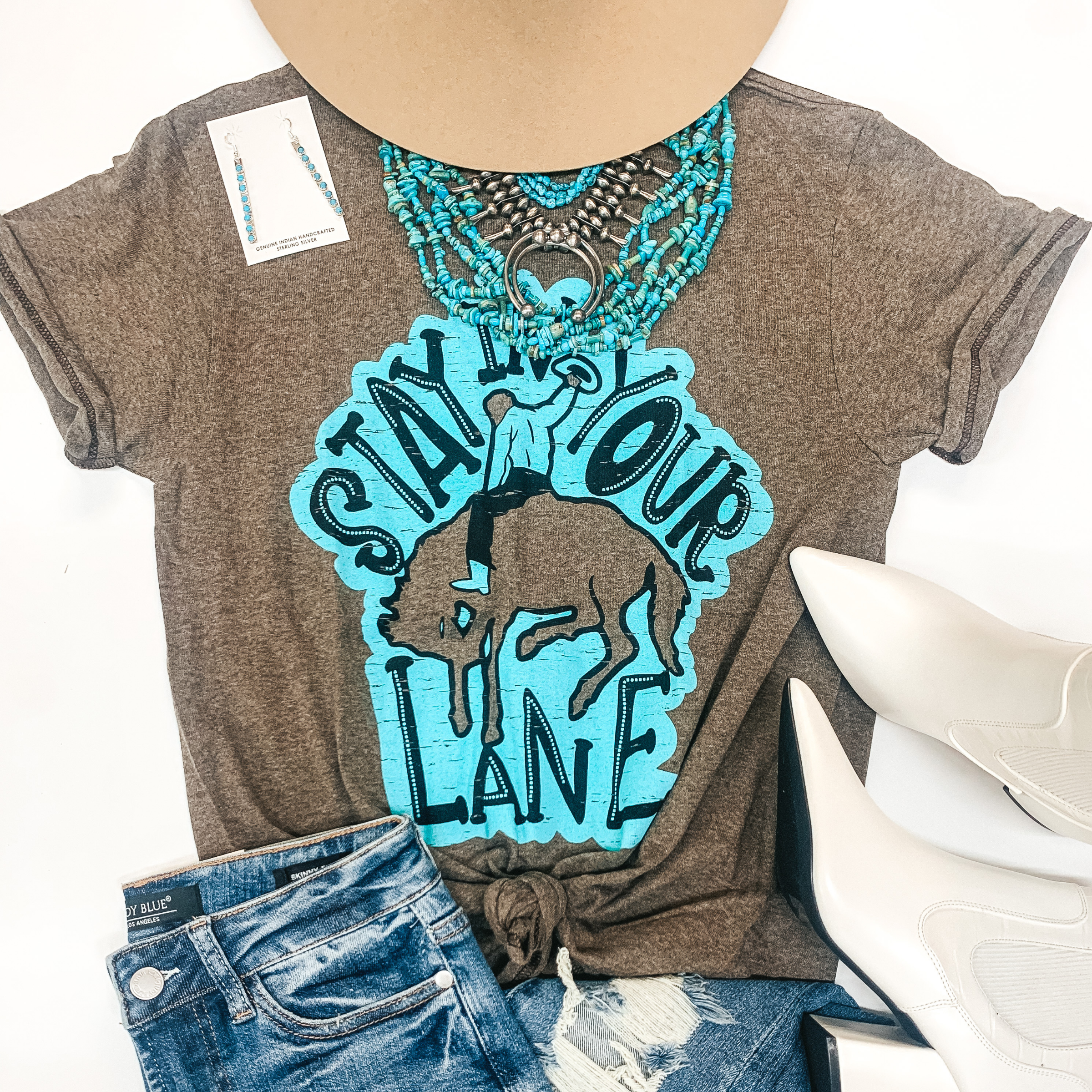 A brown tee shirt with a cowboy and a graphic that says " Stay in your lane." Pictured with turquoise jewelry, a tan hat, distressed jeans, and white booties.