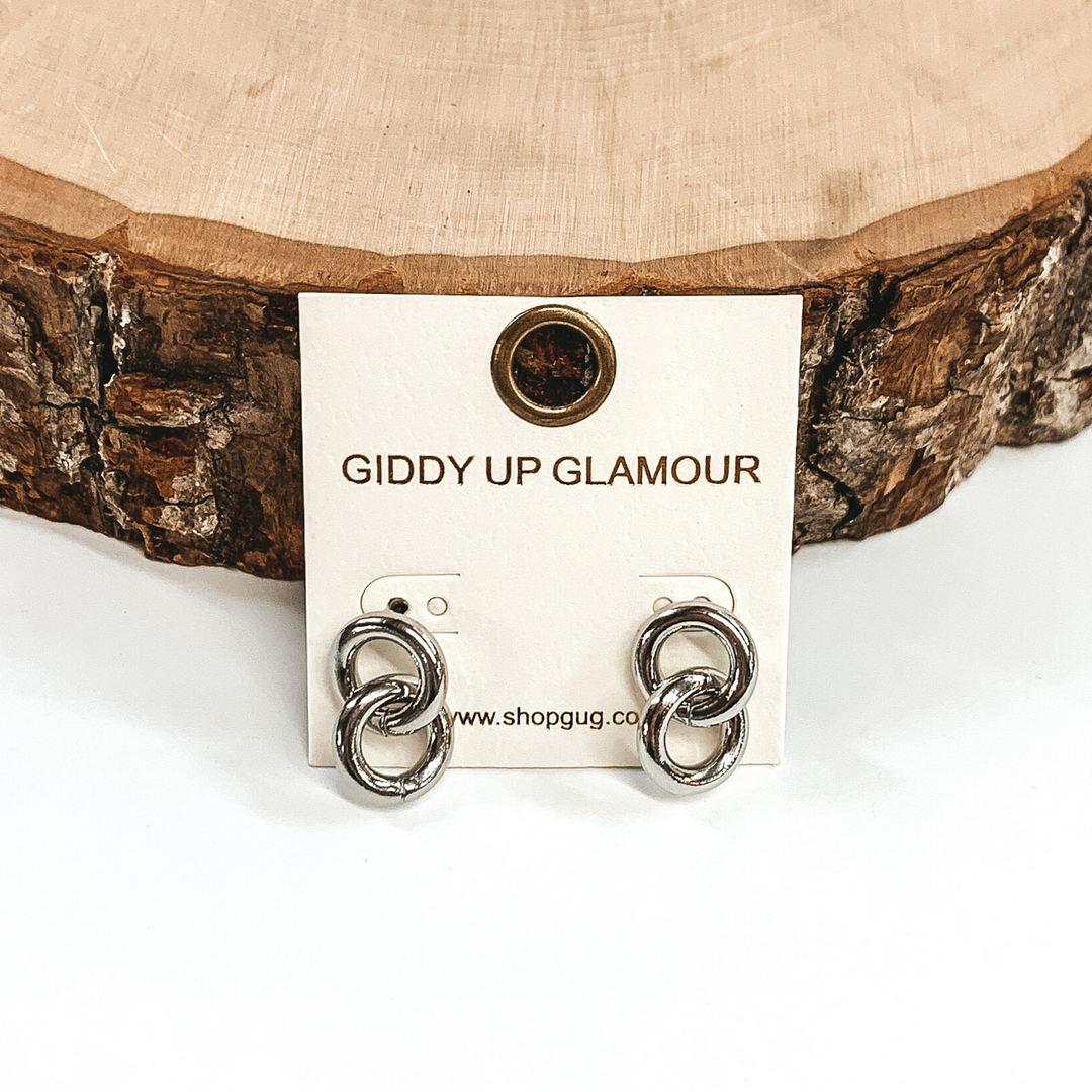 These are silver drop earrings with two circle links that are intertwined. These earrings are pictured on an ivory earring holder on a white background with a piece of wood behind the earring holder. 