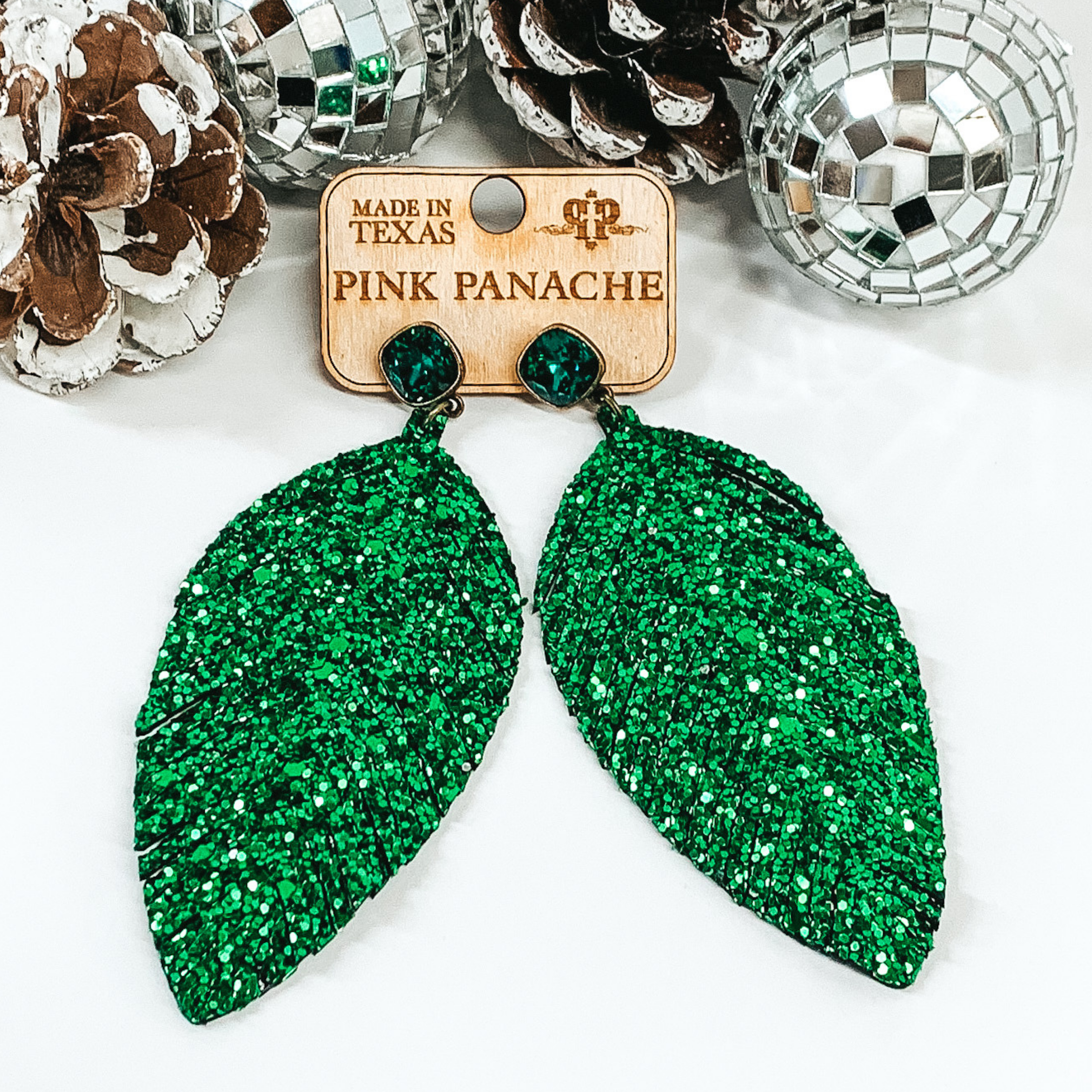 Green square crystal stud earrings with green sparkly feather pendants hanging from the bottom of the studs. Pictured on a white background with pinecones and disco balls at the top. 