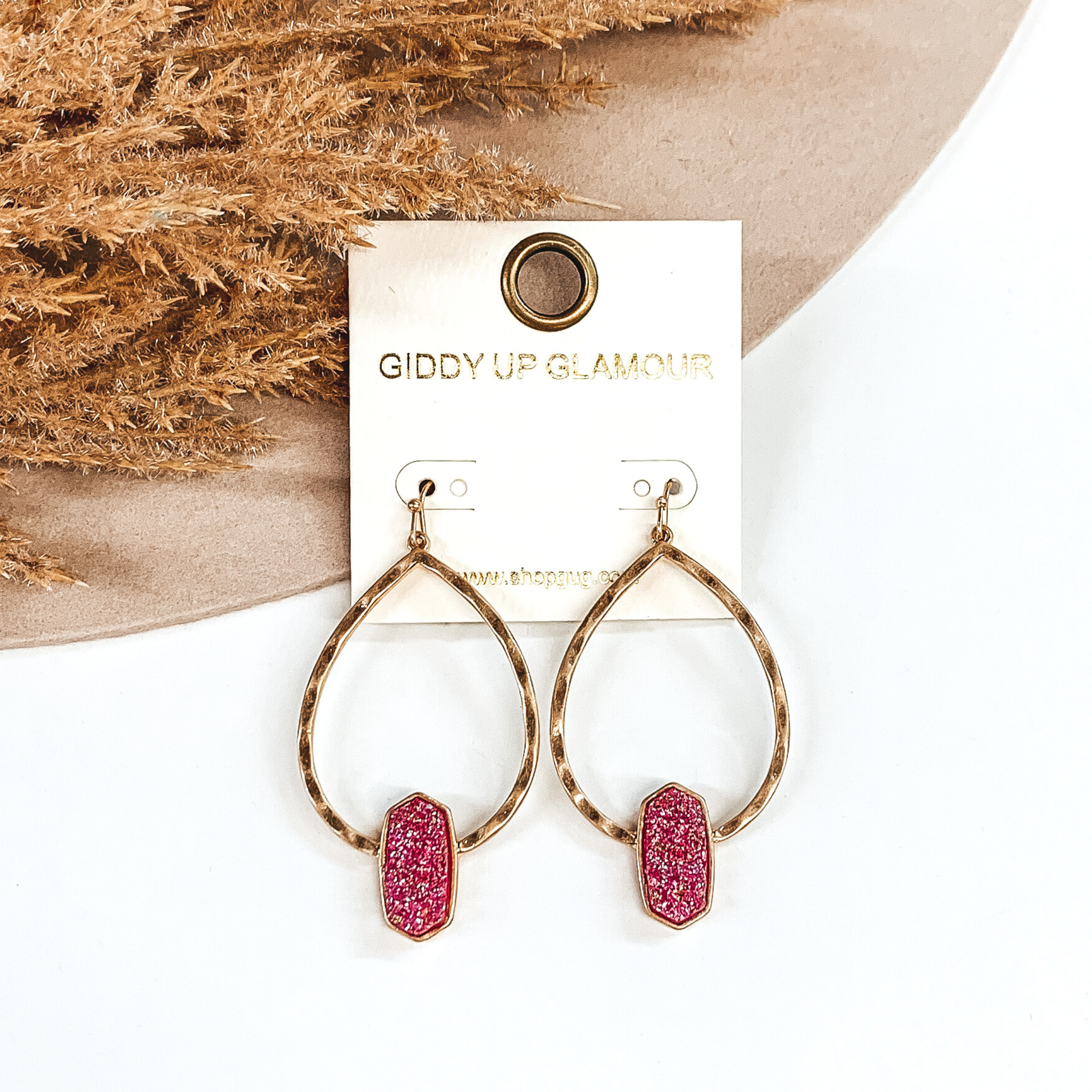 Gold hammered teardrop dangle earrings with a fuchsia druzy pendant at the bottom of the earrings. These are pictured on a white and beige background with some tan floral.