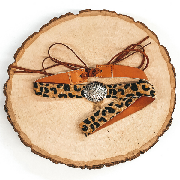 Leopard hat band with a single silver concho. It also has leather like material strands that tie it together. It is laying on a piece of wood that is pictured of a white background. 