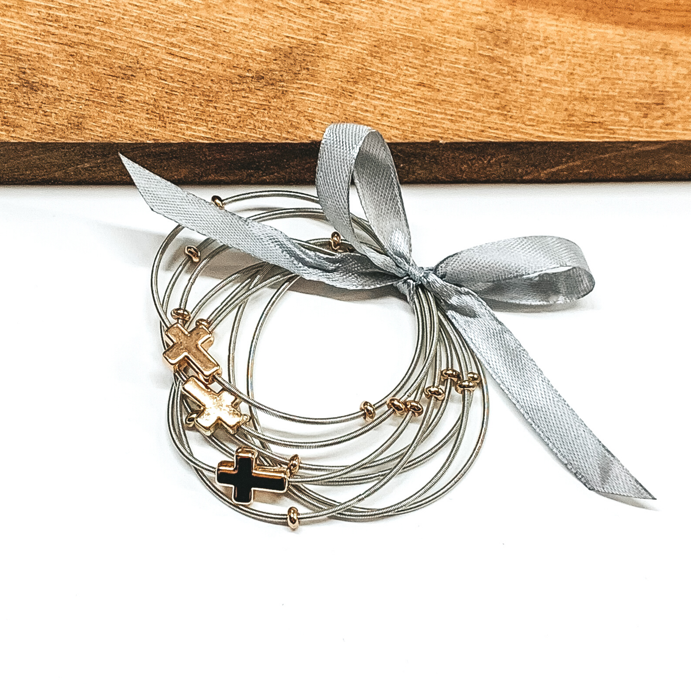 Group of silver spring wire elastic bracelet set tied together with a silver ribbon that is tied in a bow. Some of the bracelets has small gold ball beads and three bracelets have a single gold cross charm. This bracelet set is pictured laying next to a piece of wood on a white background.