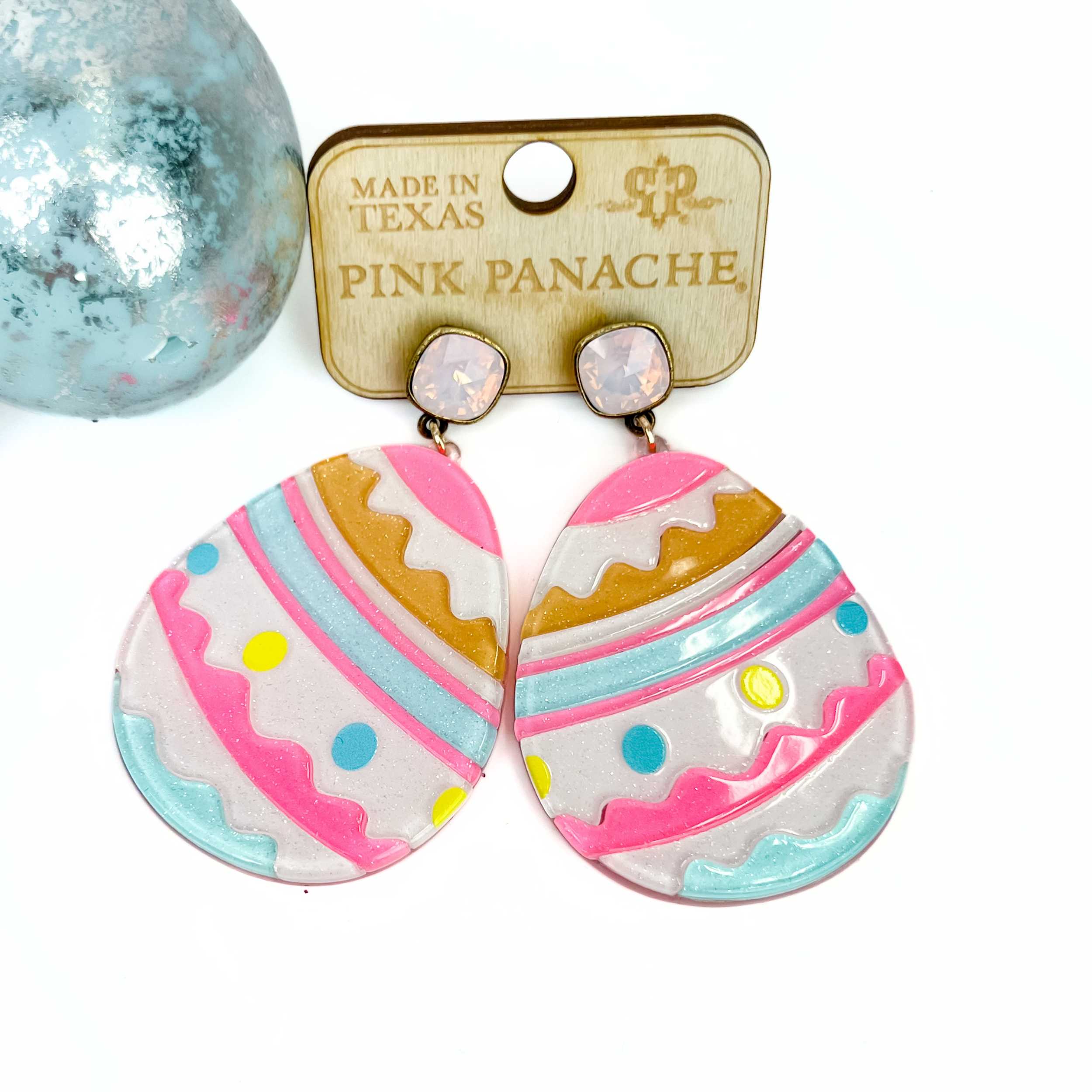 Ivory glitter acrylic easter eggs with swirly lines in pink, orange, and blue to give the appearance of an easter egg as well as neon yellow and blue dots accent the oval shaped earrings.  Attached at the top is a pink cushion cut sworvoski crystal.  Earrings are placed on a wooden earring tag that says made in Texas, Pink Panache and shows the PP logo.  Earrings have an accent of an Easter egg in the top left corner.  Earrings and egg are placed on a white background.