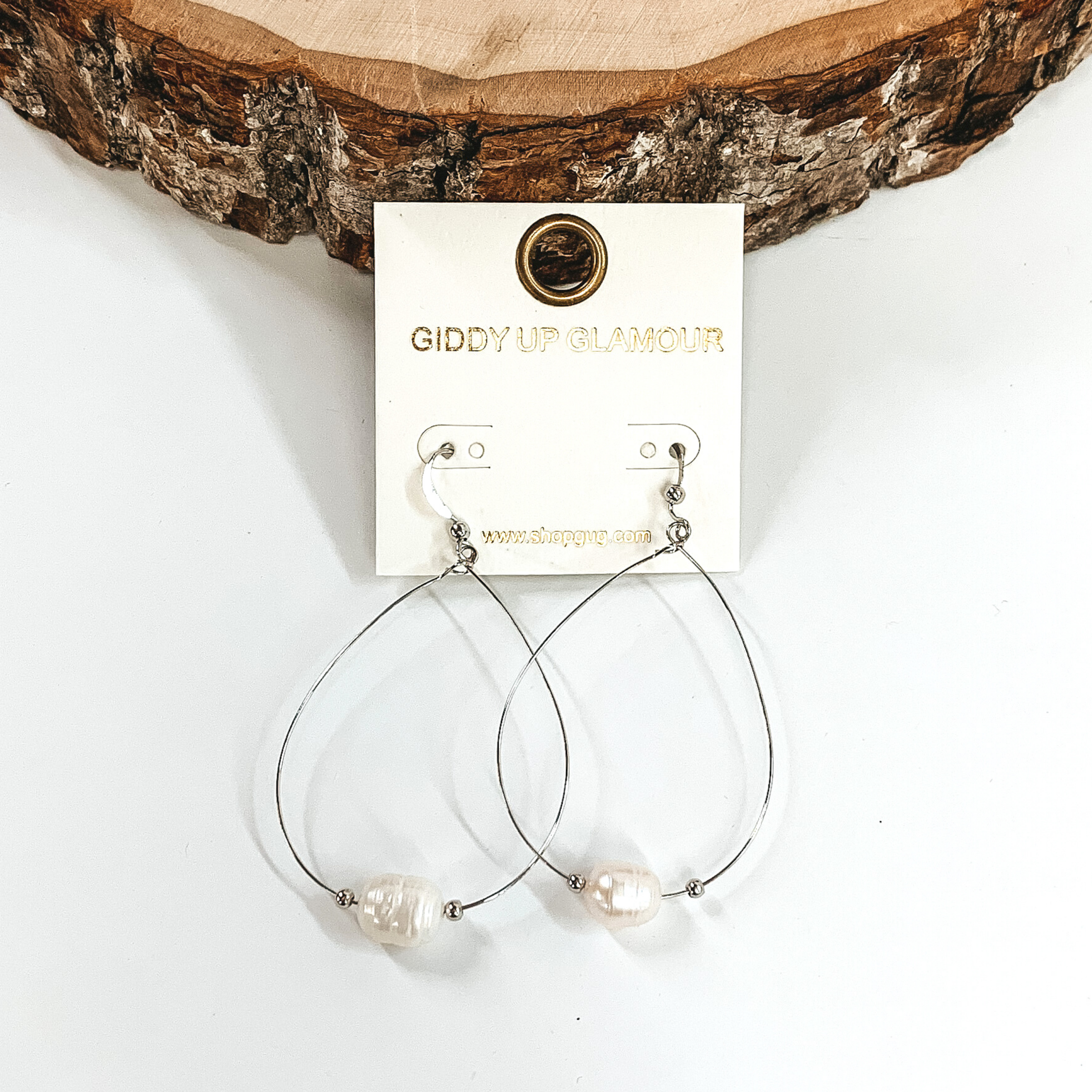Thin silver wired earrings in a teardrop shape that includes a pearl charm with two tiny silver beads on either side of the pearl. These earrings are pictured on a white background with a piece of wood at the top. 