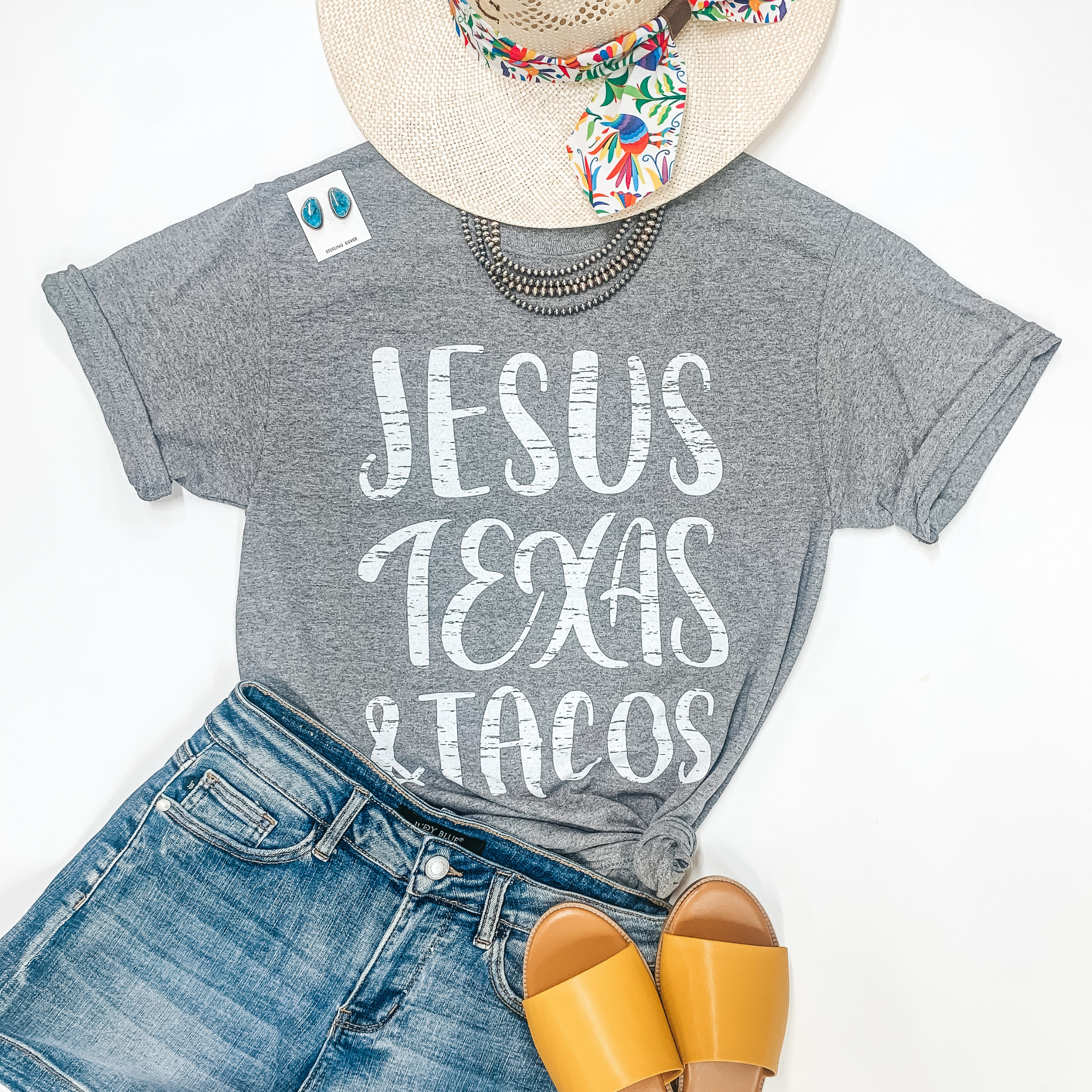 A grey tee shirt with "Jesus Texas & Tacos." Pictured with a straw hat, genuine Navajo jewelry, yellow sandals, and denim shorts.