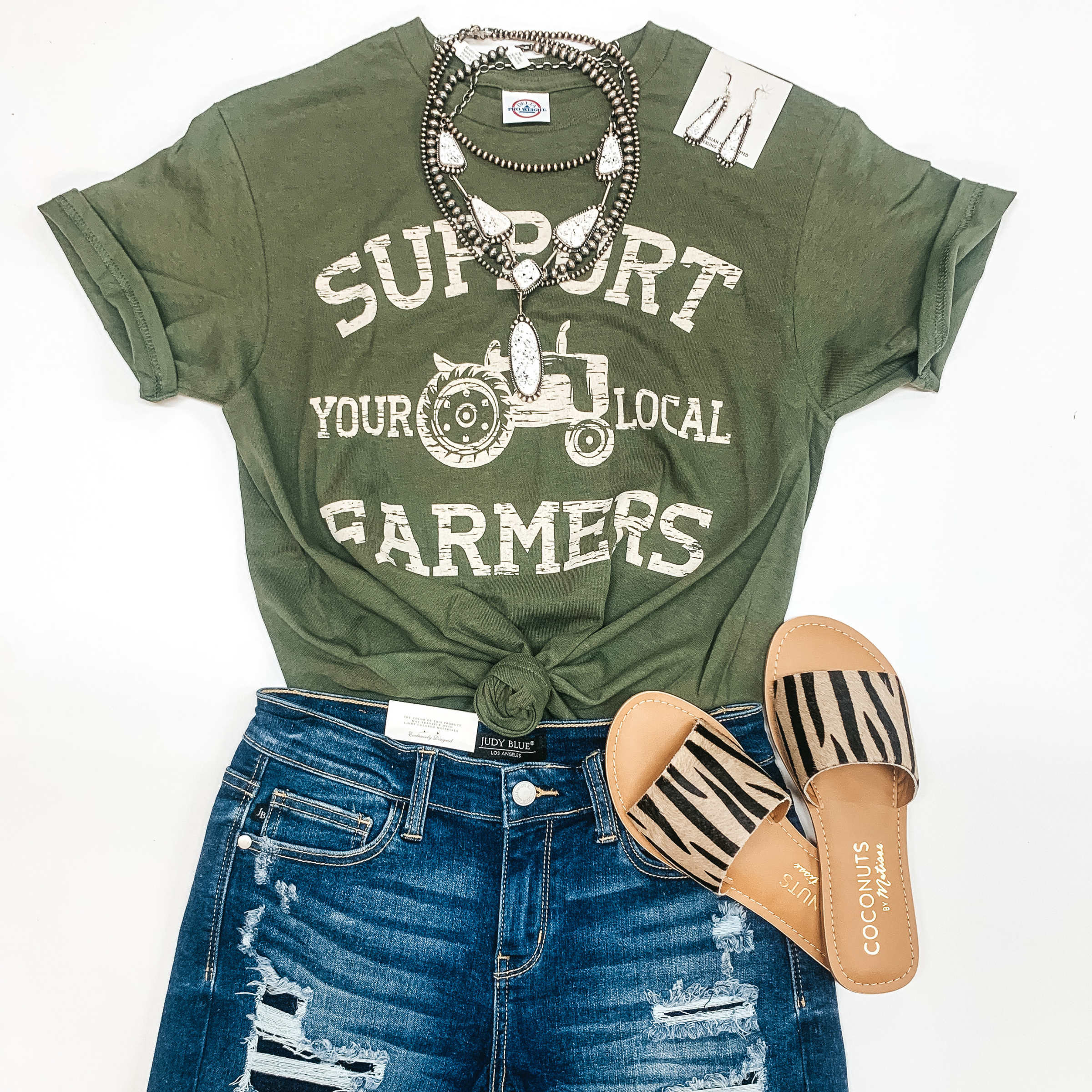 An olive green tee shirt with a tractor that says "Support your local farmers." Pictured with genuine Navajo jewelry, denim shorts, and sandals.