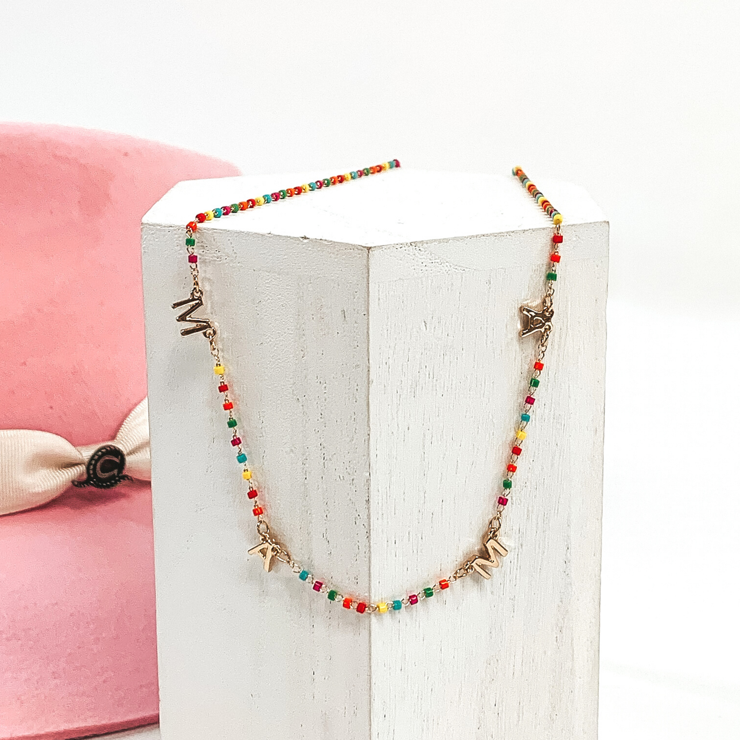 This is a multicolored beaded necklace with gold letter charms that spell out "MAMA." This necklace is pictured on a white block on a white background with a pink hat in the background.