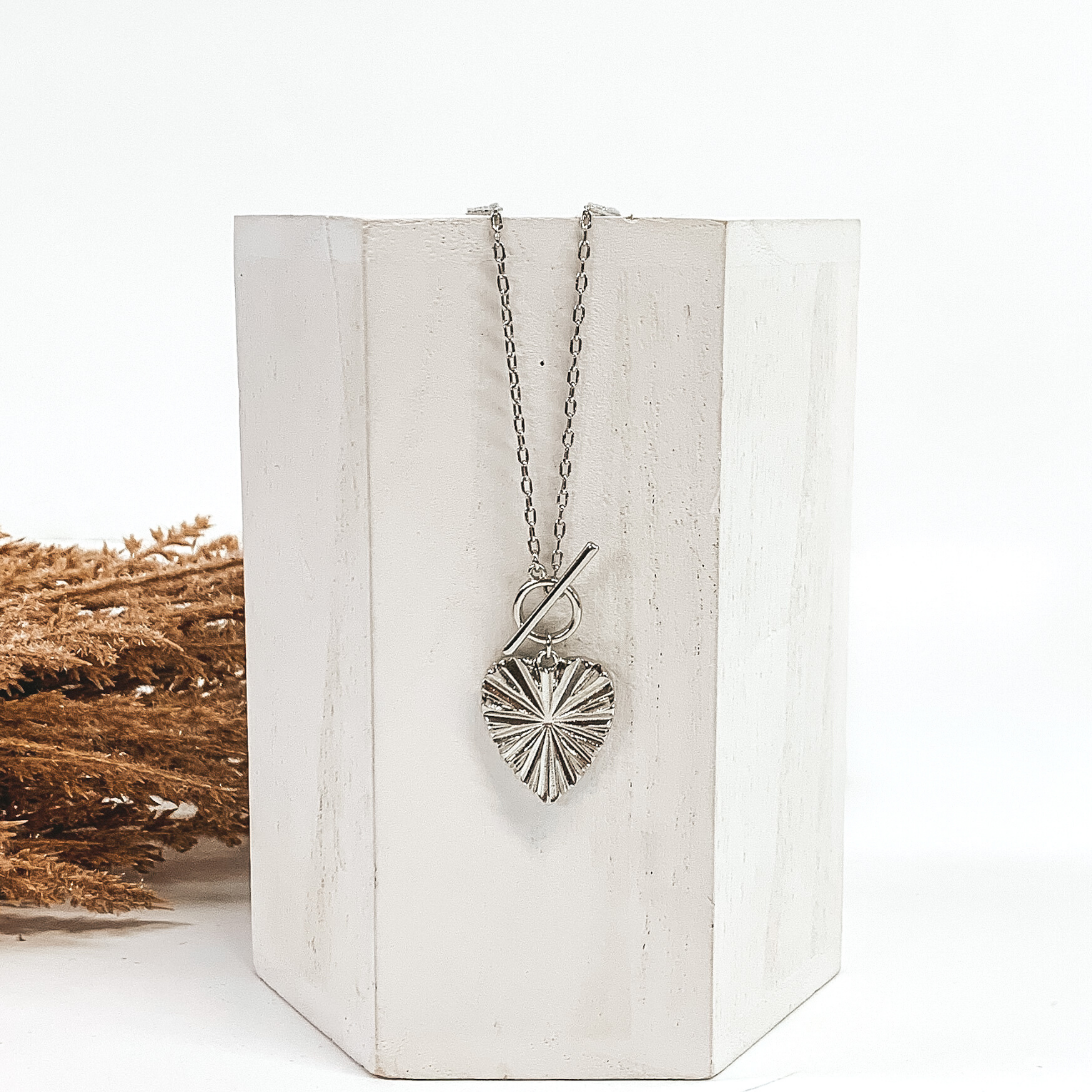 This is a silver necklace with a shell textured heart pendant and a front toggle clasp. This necklace is pictured laying on a white block and on a white background with tan floral on the left side of the block. 