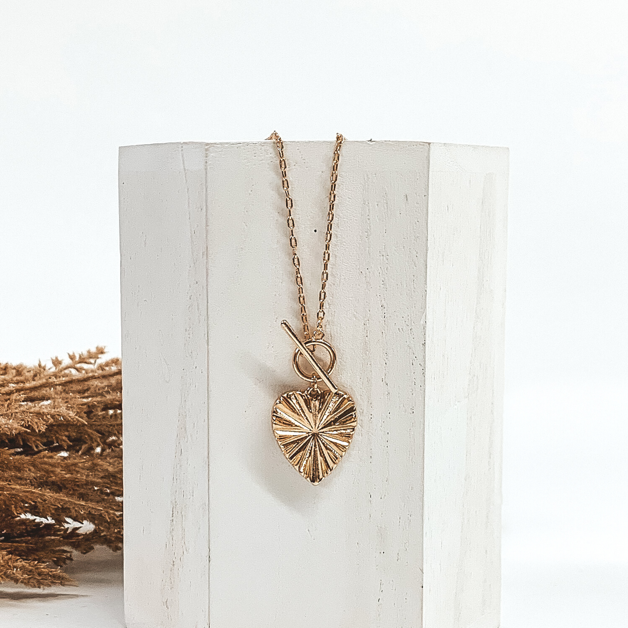 This is a gold necklace with a shell textured heart pendant and a front toggle clasp. This necklace is pictured laying on a white block and on a white background with tan floral on the left side of the block.