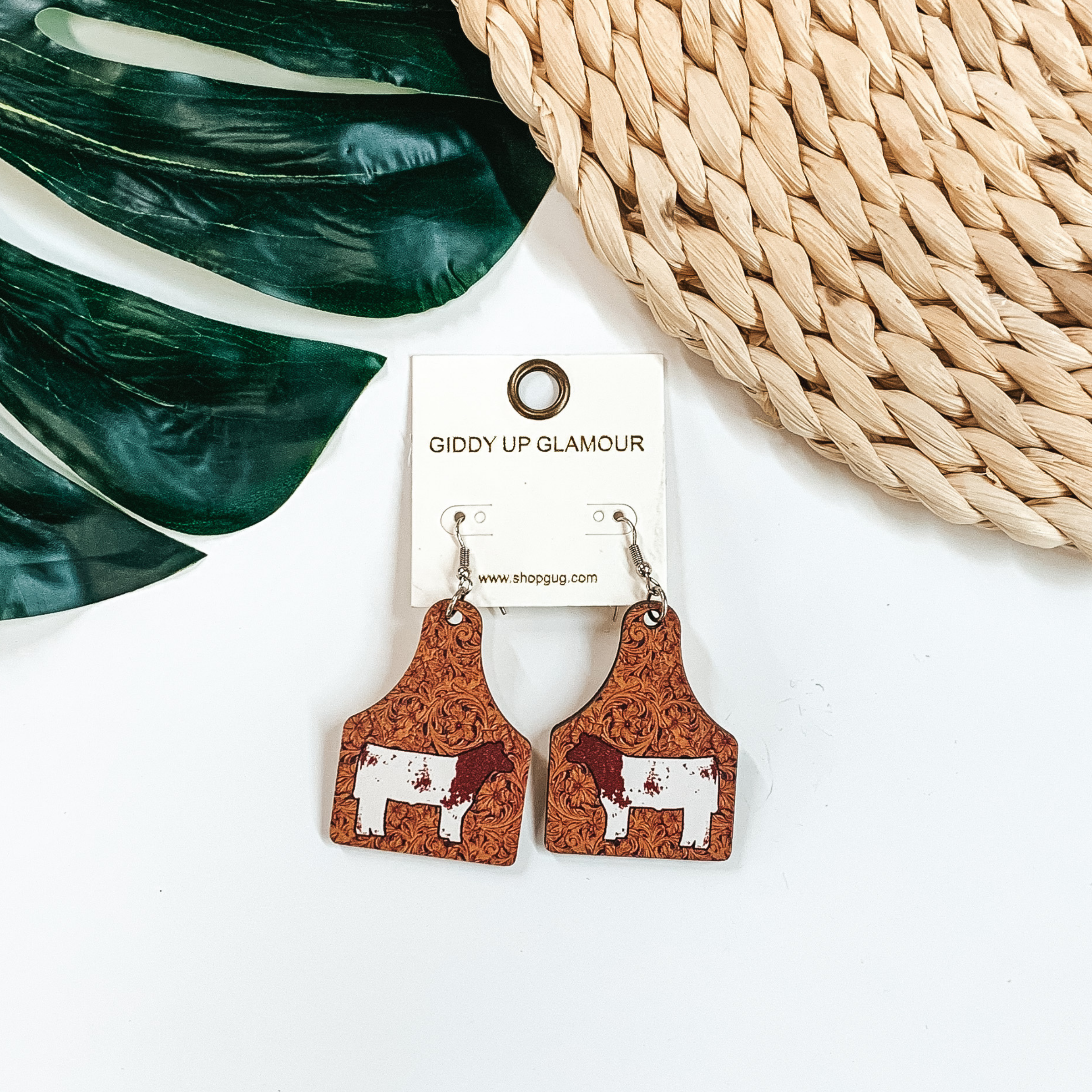 Cattle tag drop earrings with a tooled leather print. In the center of the earrings is a brown and white cow. These earrings are pictured on a white background with a green leaf and tan basket weave material at the top of the picture. 
