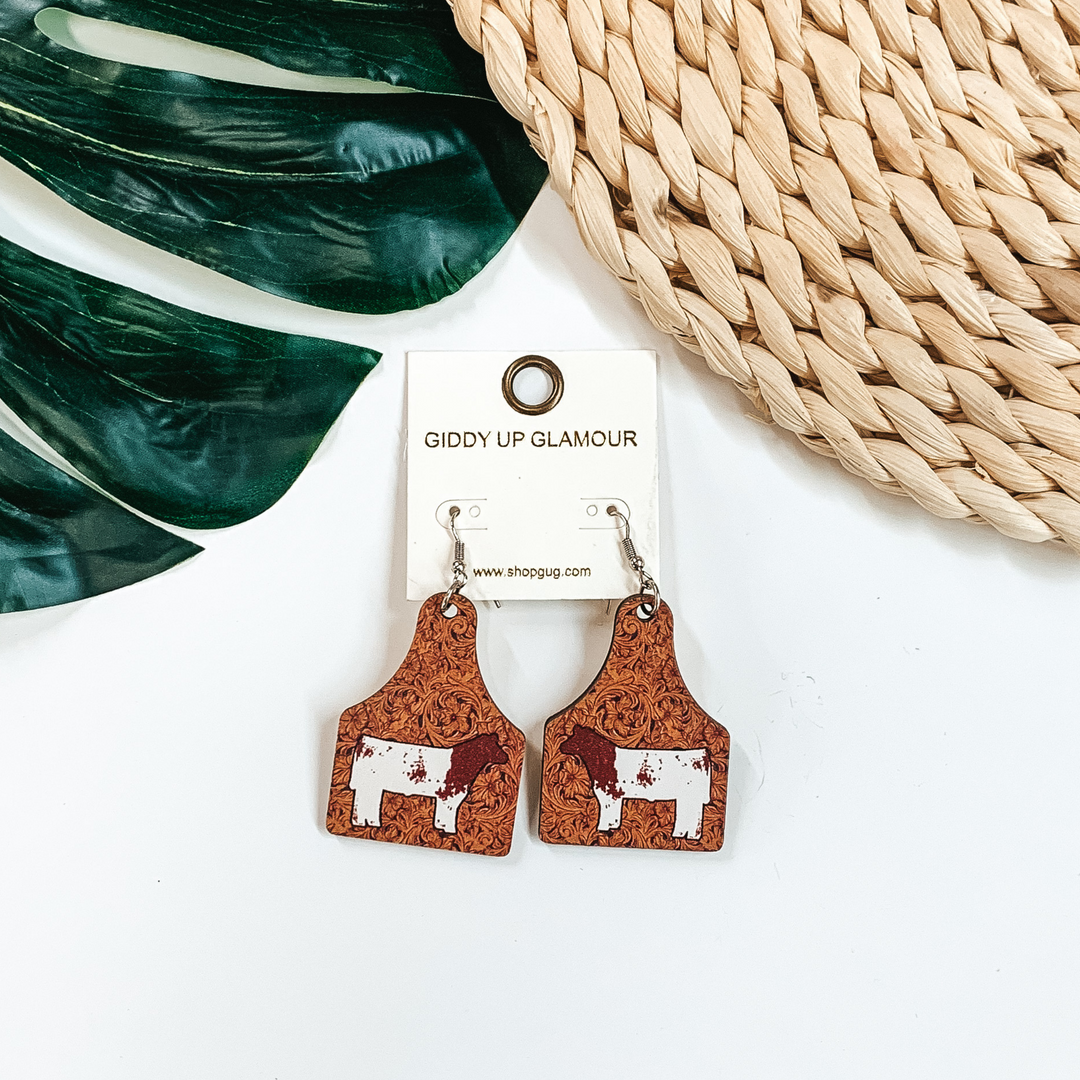 Cattle tag drop earrings with a tooled leather print. In the center of the earrings is a brown and white cow. These earrings are pictured on a white background with a green leaf and tan basket weave material at the top of the picture. 