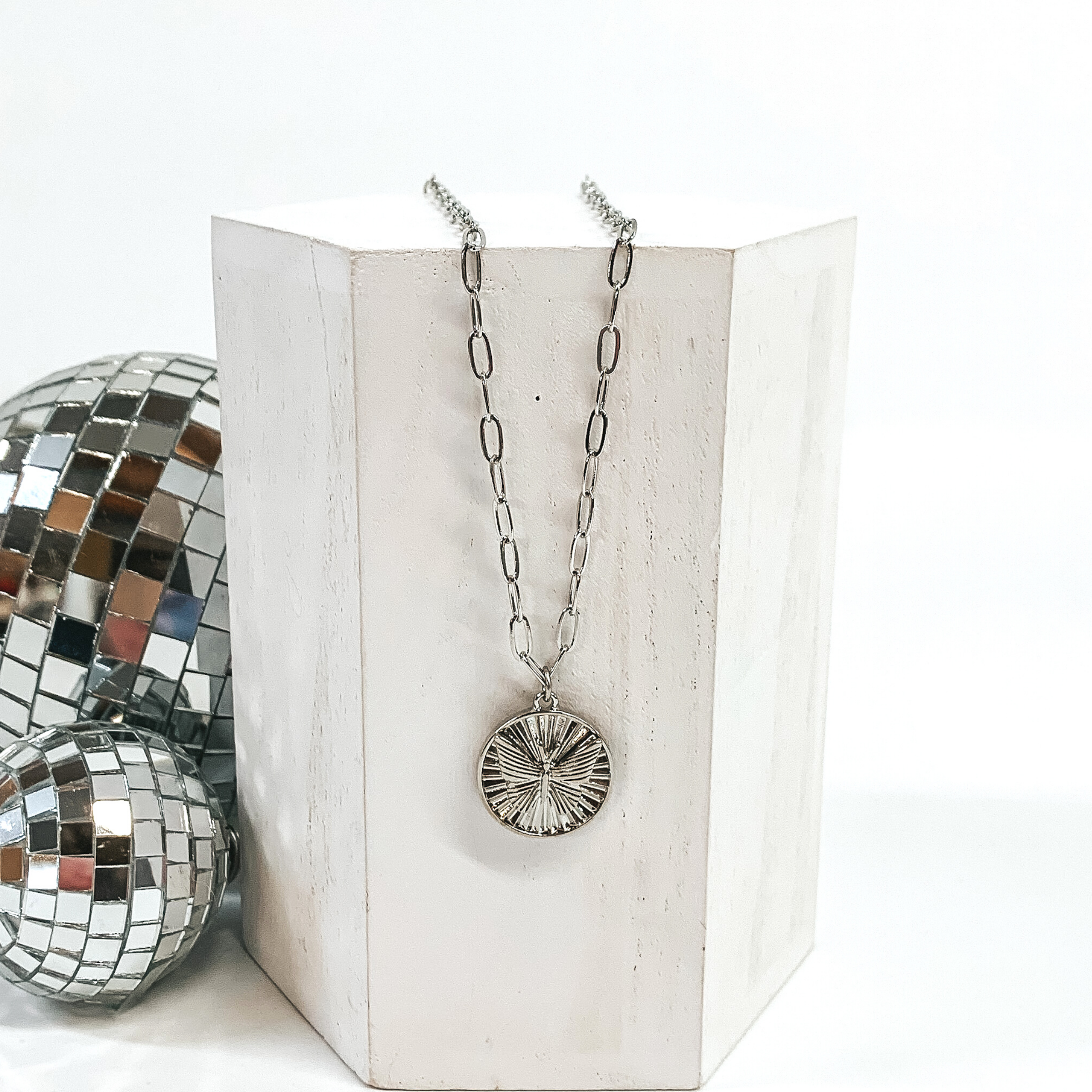 This is a silver necklace with a shell textured circle pendant and a butterfly charm on the front of the circle pendant. This necklace is pictured laying on a white block and on a white background with disco balls on the left side of the block.