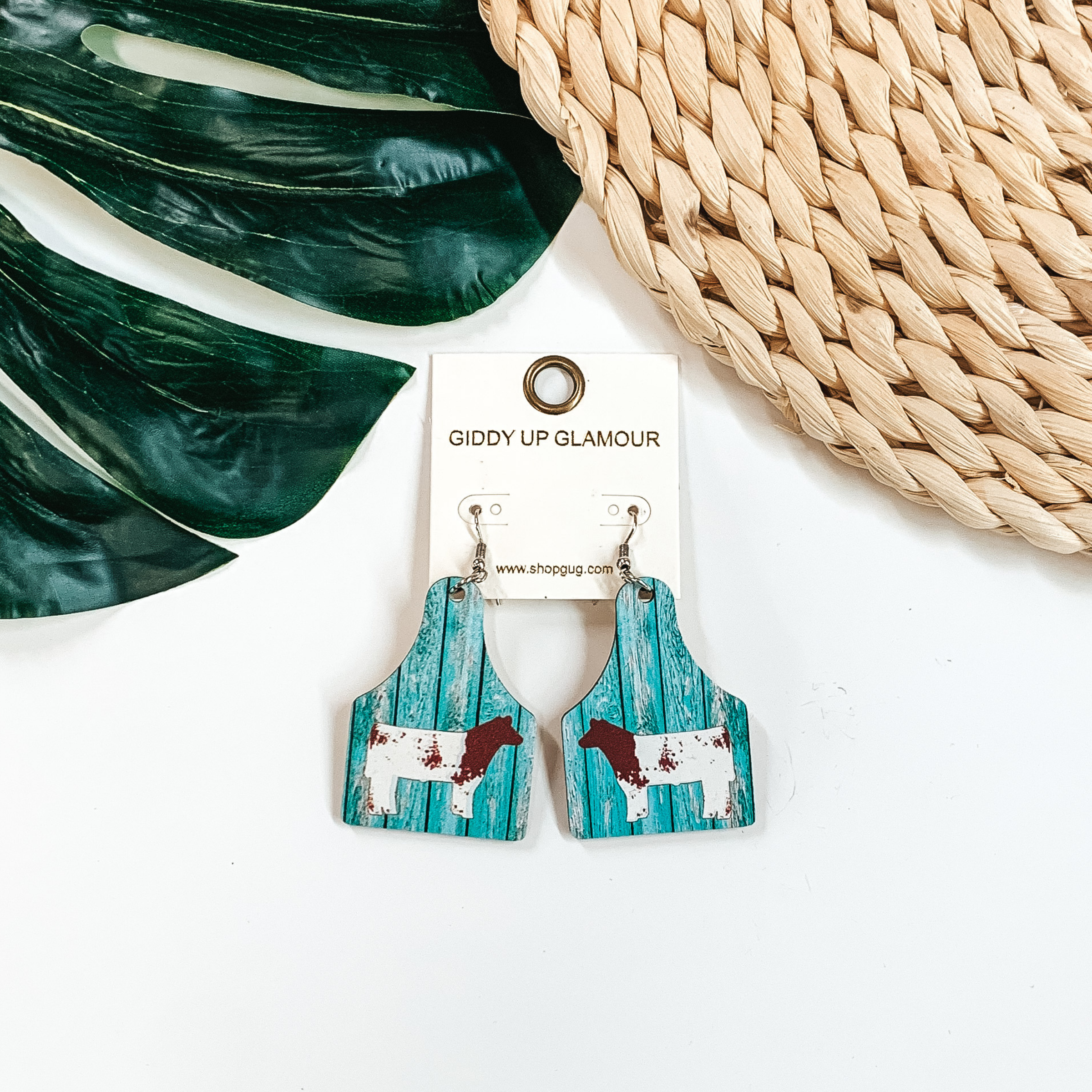 Cattle tag drop earrings with a turquoise wood print. In the center of the earrings is a brown and white cow. These earrings are pictured on a white background with a green leaf and tan basket weave material at the top of the picture. 