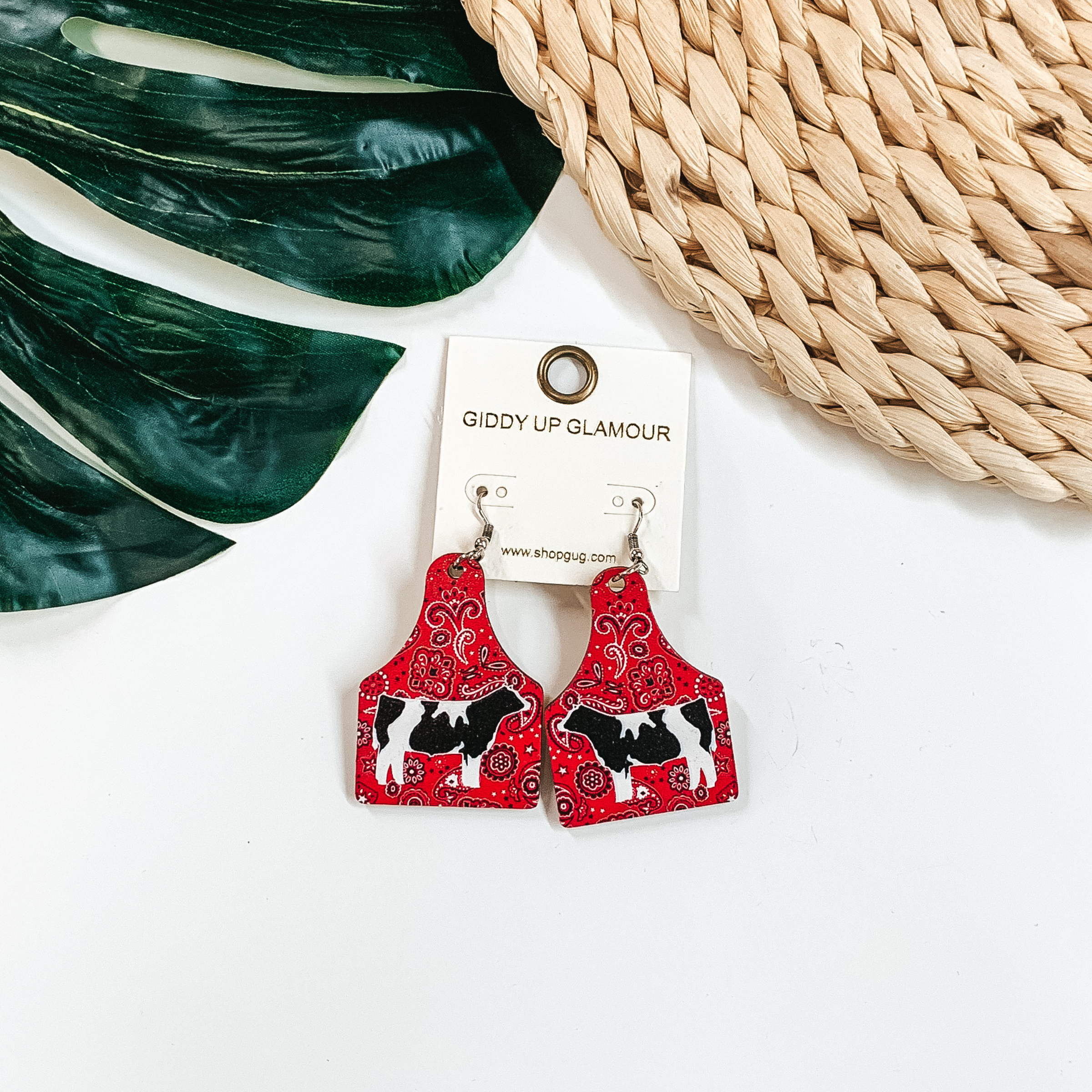 Cattle tag drop earrings in a red paisley print. In the center of these earrings there is a picture of a black and whtie cow. These earrings are pictured on a white background with a green leaf. 