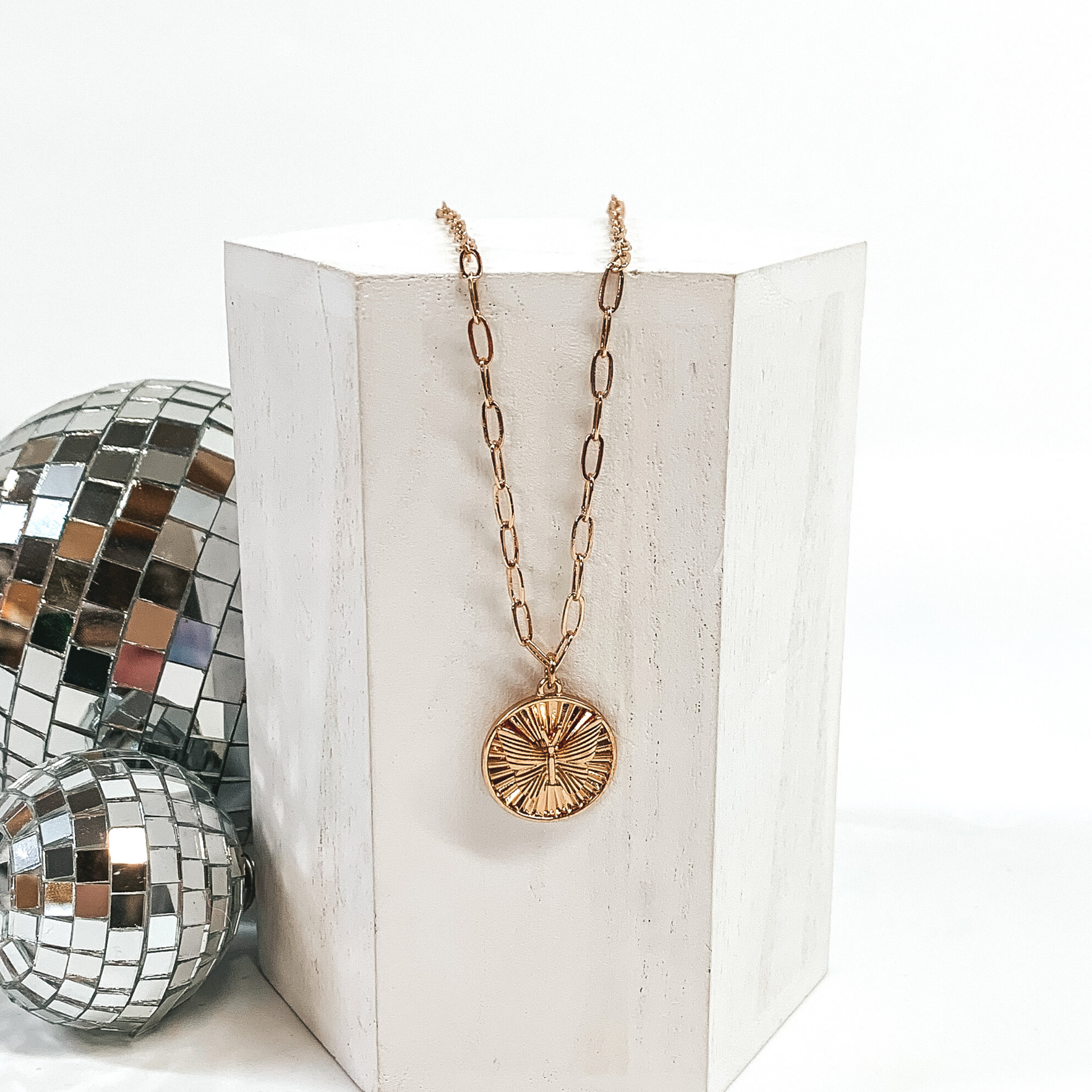 This is a gold necklace with a shell textured circle pendant and a butterfly charm on the front of the circle pendant. This necklace is pictured laying on a white block and on a white background with disco balls on the left side of the block.