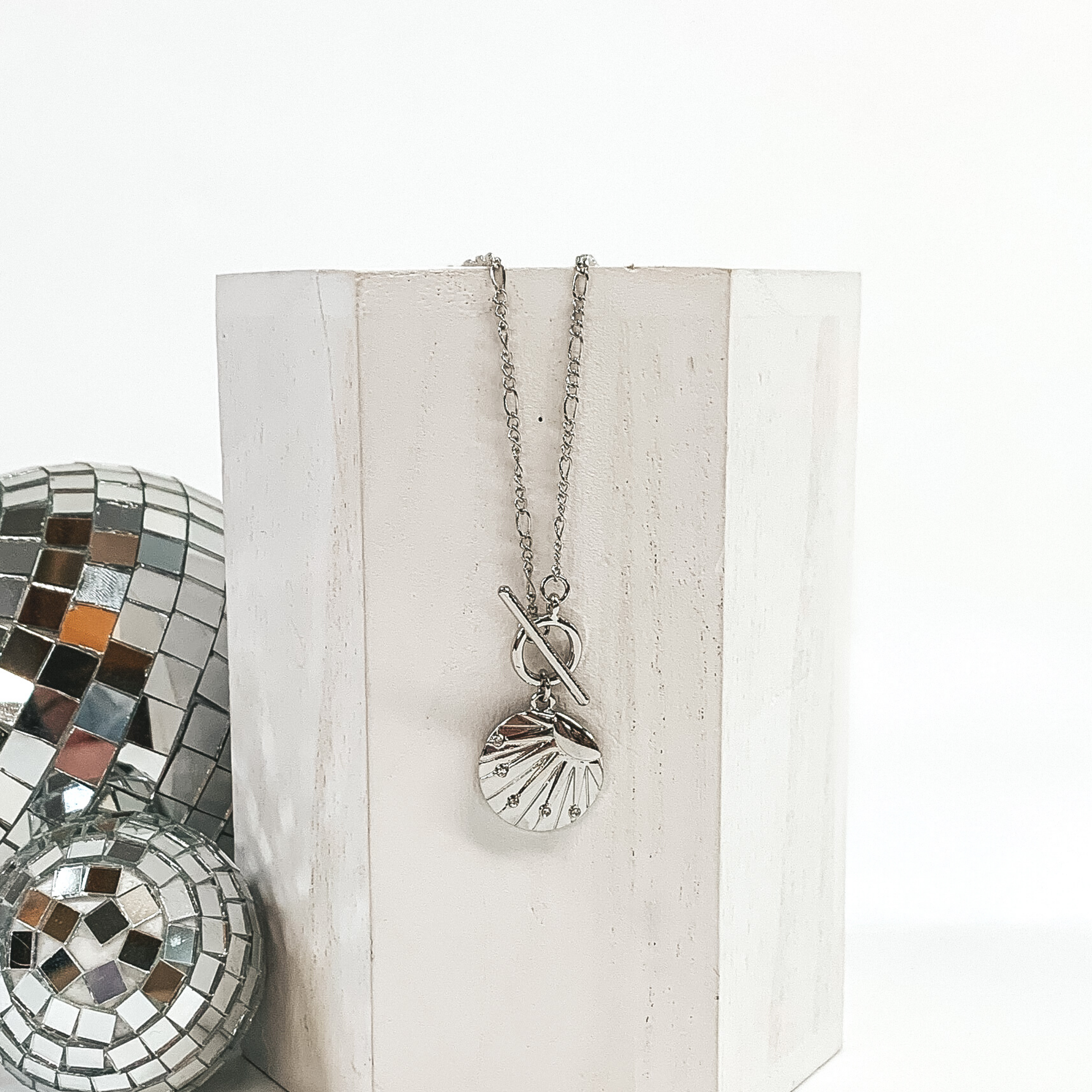 This is a silver necklace with a shell textured circle pendant with a sun design on it with a front toggle clasp. This necklace is pictured laying on a white block and on a white background with disco balls on the left side of the block.