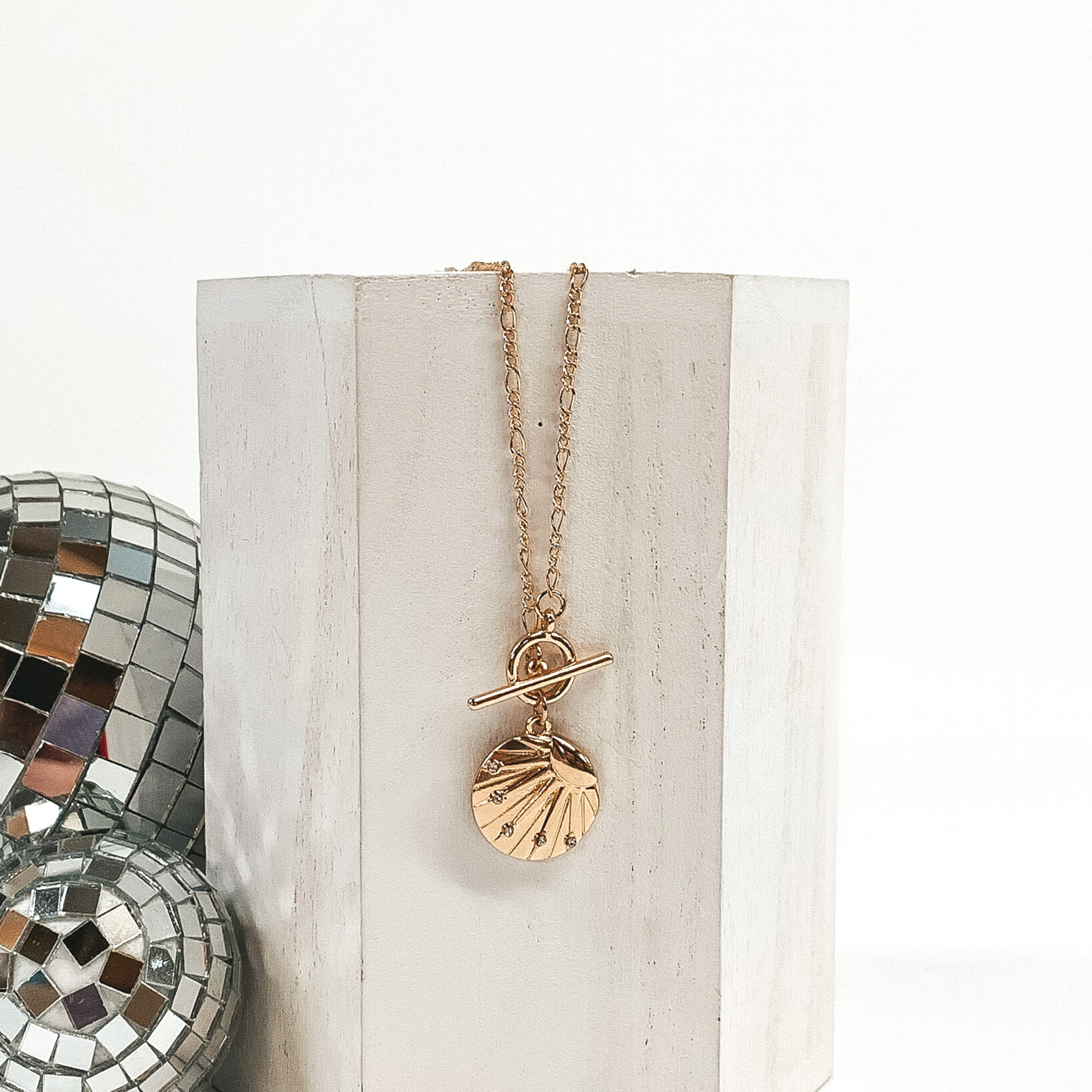 This is a gold necklace with a shell textured circle pendant with a sun design on it with a front toggle clasp. This necklace is pictured laying on a white block and on a white background with disco balls on the left side of the block.