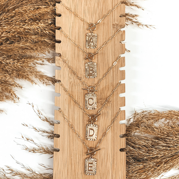 There are five gold initial necklaces on a light tan necklace holder stacked one on top of the other. The gold rectangle pedants on each necklace has an engraved letter in the center. There is also a front toggle clasp. This is pictured on a white background with tan floral at the top and bottom of the picture. 