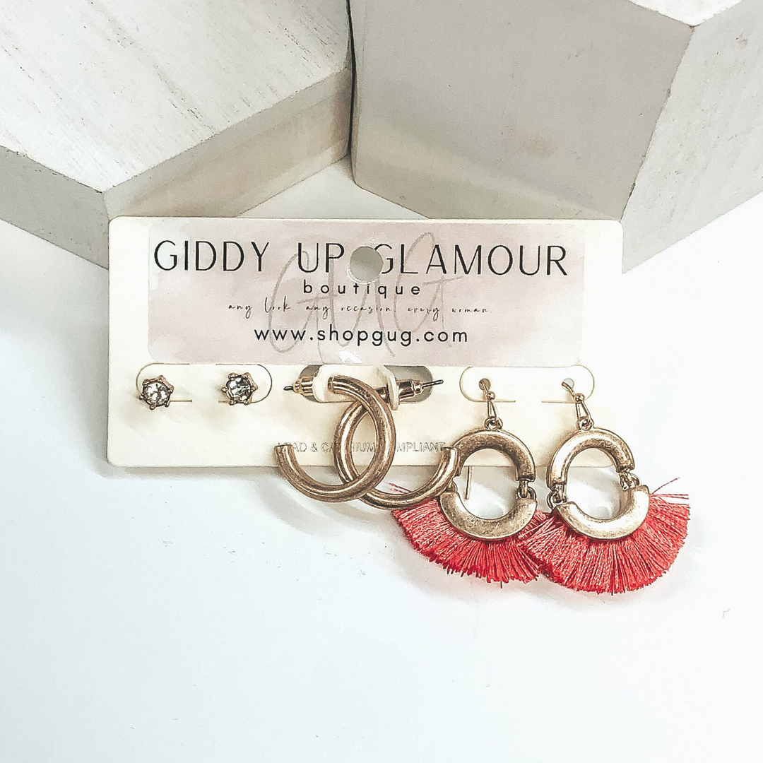 This is a three piece earring set. You have a pair of gold studs with clear crystals, a pair of gold hoops, and gold oval dangle earrings with coral tassels. These are pictured on an earrings holder on a white background.