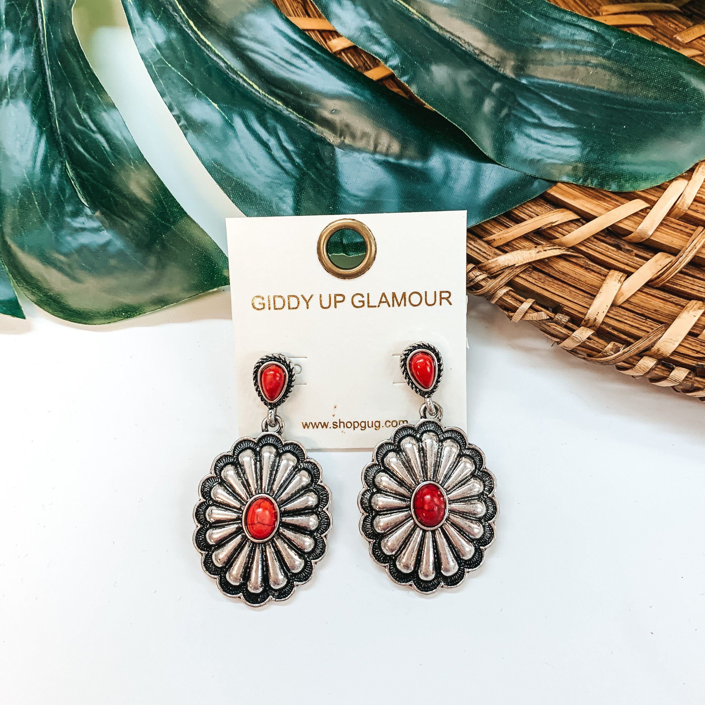 A pair of silver concho earrings with red stones. These earrings are pictured on a white background with a basket and palm leaf.