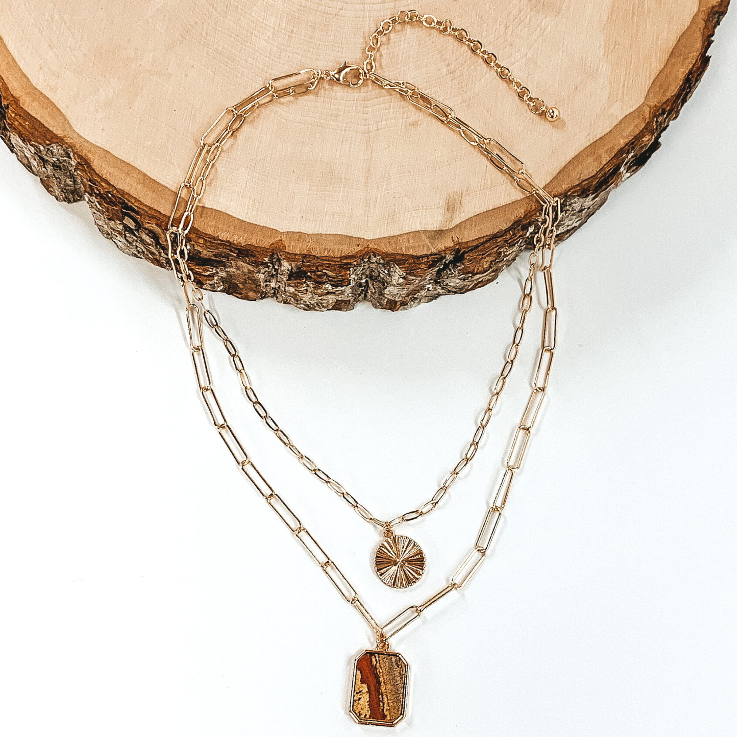 Double layered paperclip chain necklace in gold. The shorter strand has a gold circle pendant and the longer strand has a tan stone pendant in a long octagon shape. This necklace is half laying on a piece of wood and is pictured on a white background.