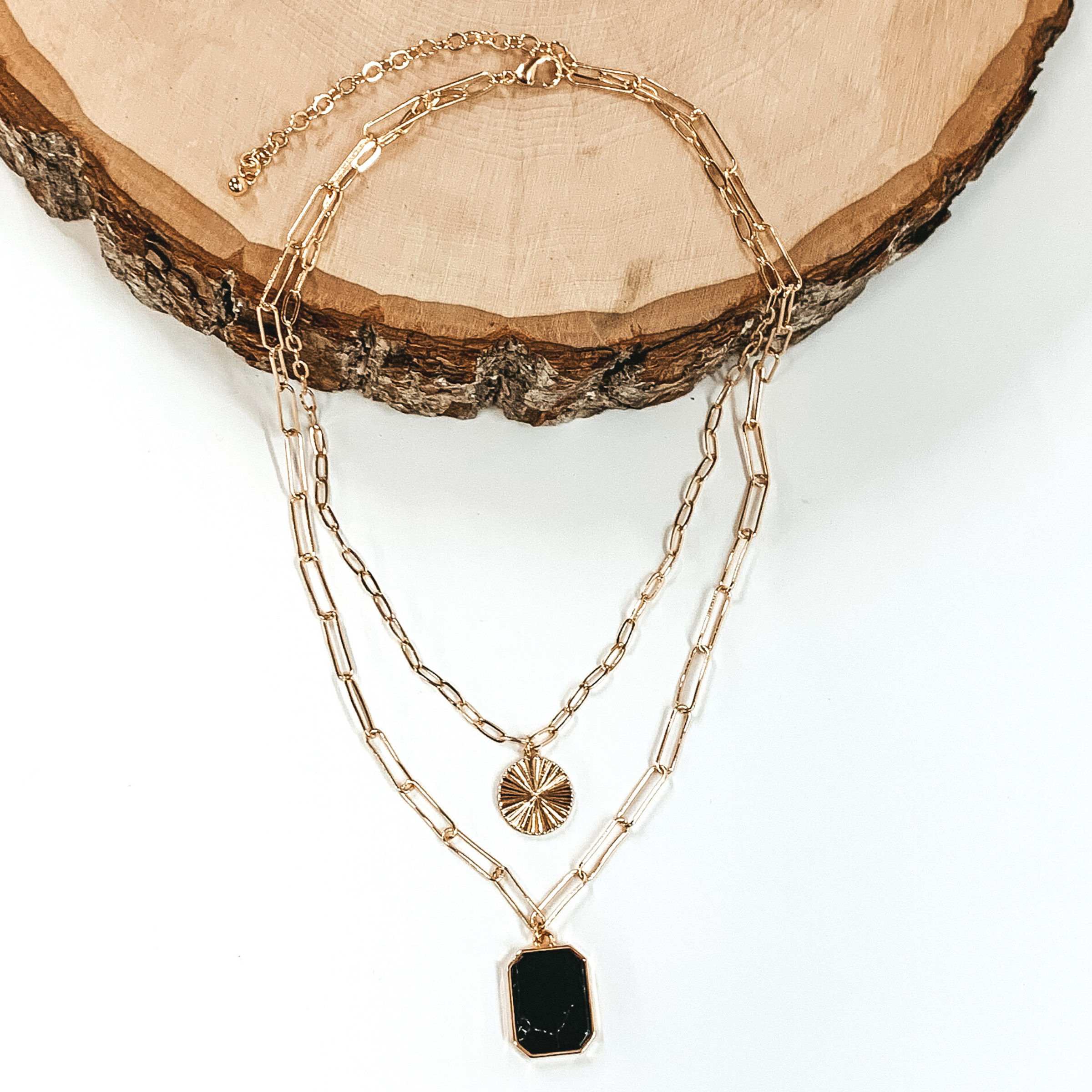 Double layered paperclip chain necklace in gold. The shorter strand has a gold circle pendant and the longer strand has a black stone pendant in a long octagon shape. This necklace is half laying on a piece of wood and is pictured on a white background.