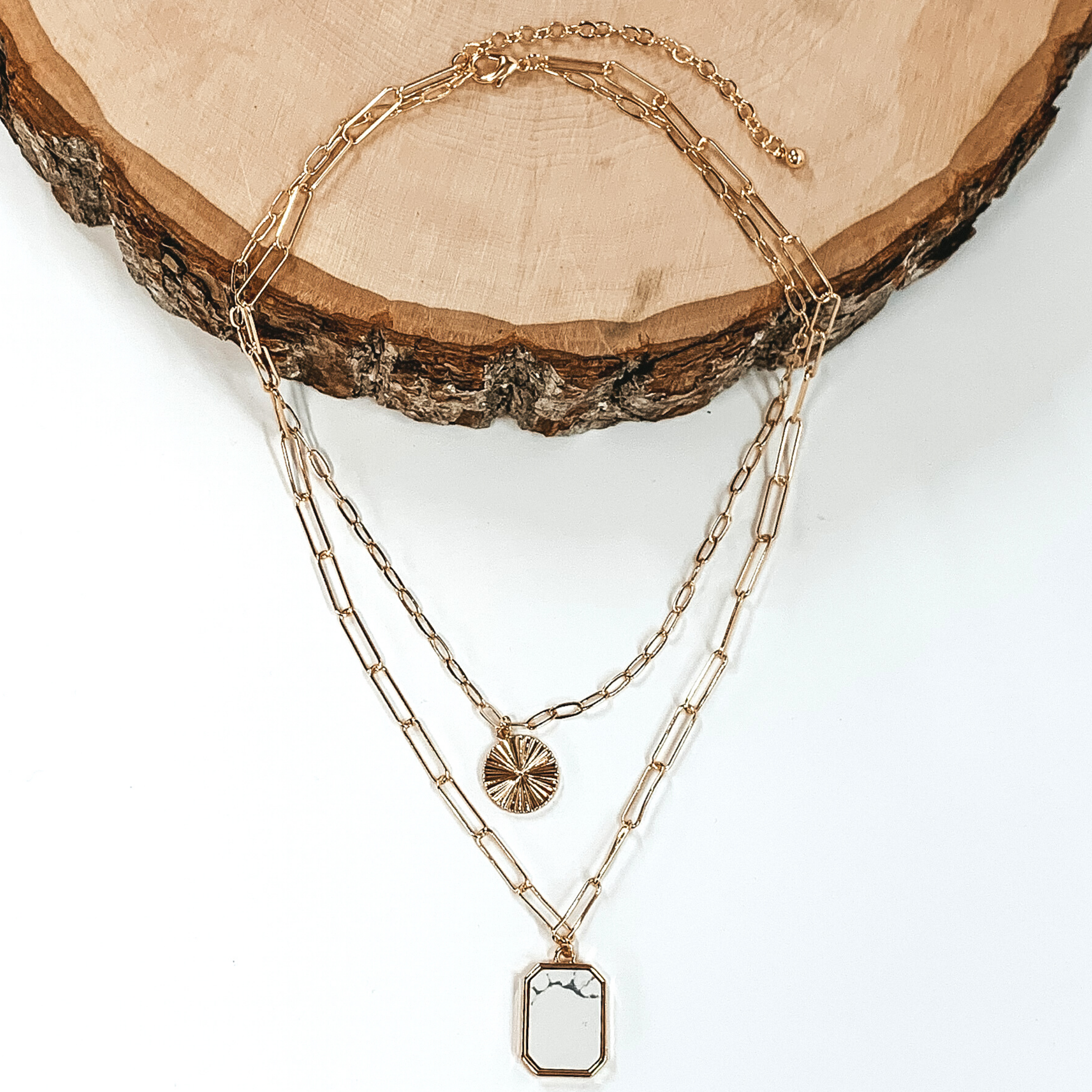 Double layered paperclip chain necklace in gold. The shorter strand has a gold circle pendant and the longer strand has a white stone pendant in a long octagon shape. This necklace is half laying on a piece of wood and is pictured on a white background.
