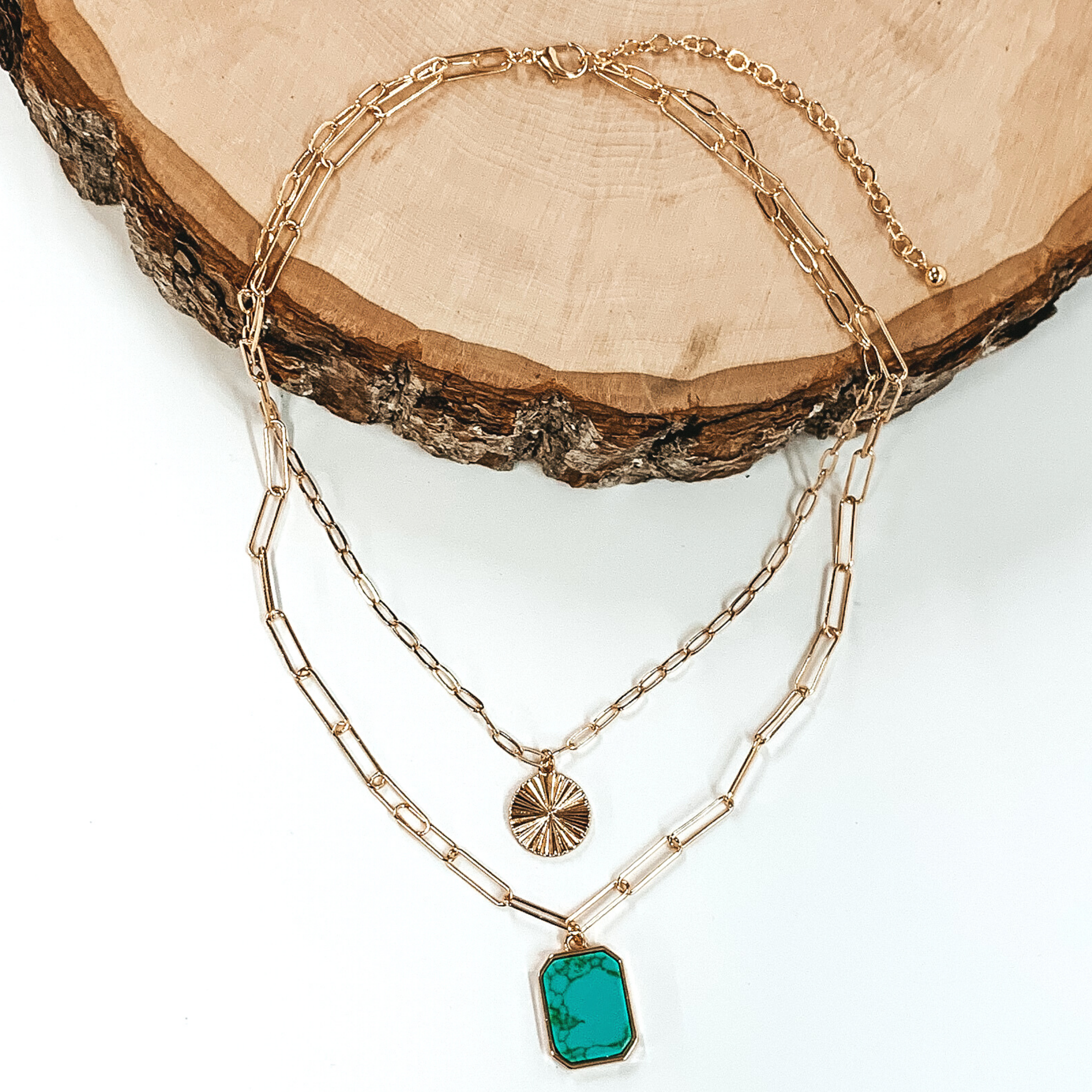 Double layered paperclip chain necklace in gold. The shorter strand has a gold circle pendant and the longer strand has a turquoise stone pendant in a long octagon shape. This necklace is half laying on a piece of wood and is pictured on a white background.