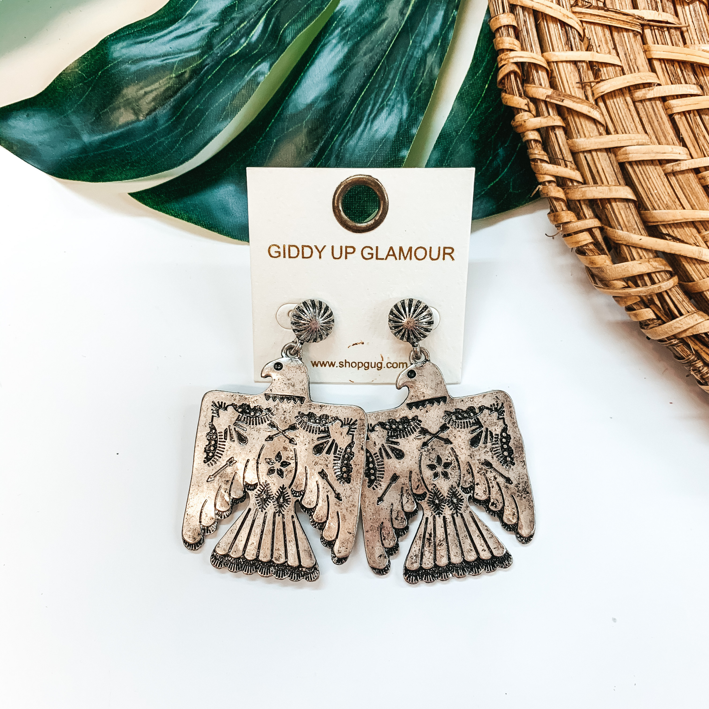 Silver, thunderbird drop earrings on a silver concho post back. These earrings are pictured on a white background in front of a green leaf. 