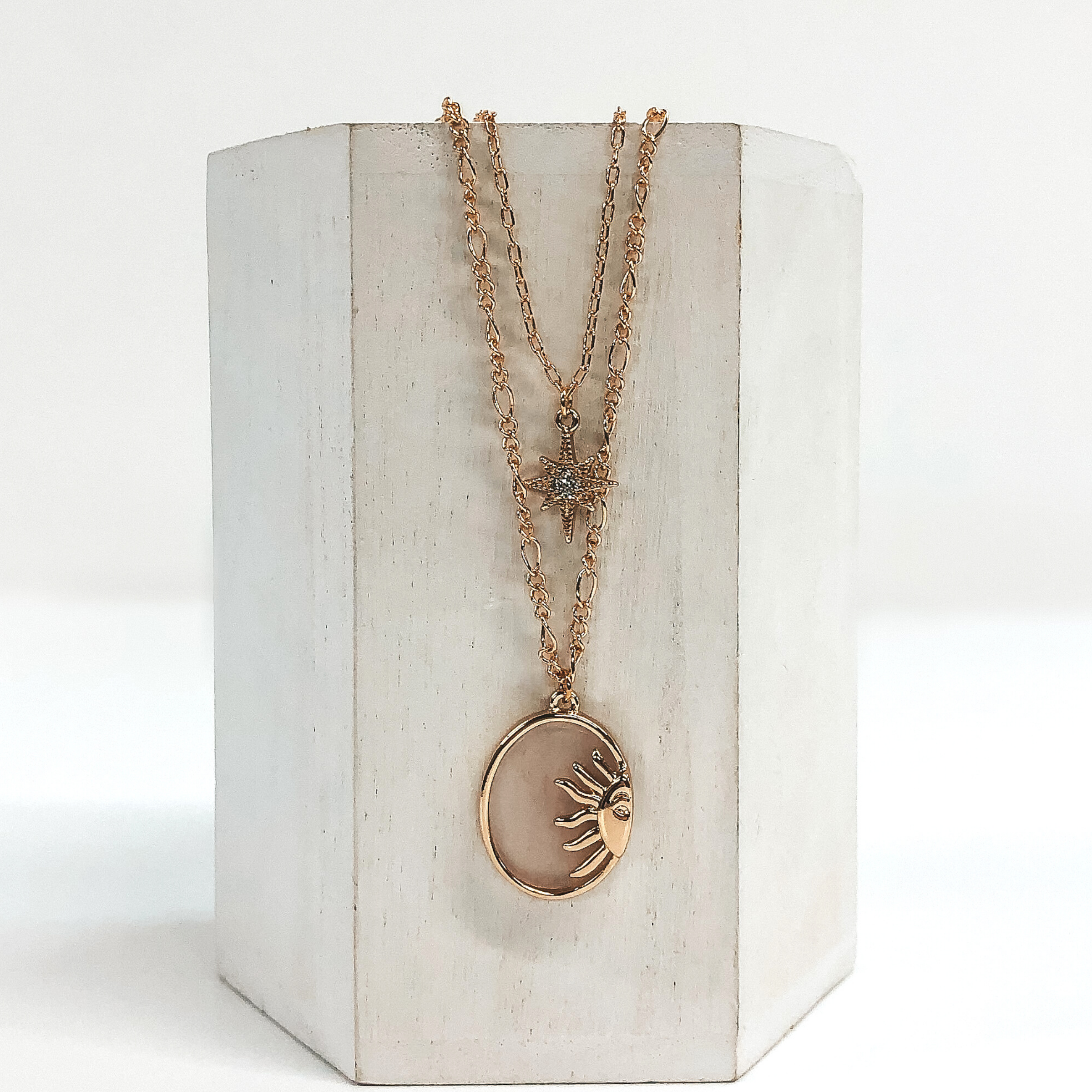 Double layered chain necklace in gold. The shorter strand has a gold star pendant with a center clear crystal and the longer strand has an oval opaque rose stone pendant with a half sun gold charm. This necklace is pictured hanging on a white block on a white background. 