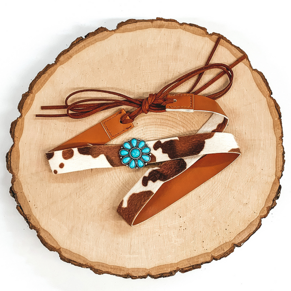 Brown cow print hat band with a single turquoise stone cluster concho. It also has leather like material strands that tie it together. It is laying on a piece of wood that is pictured of a white background.