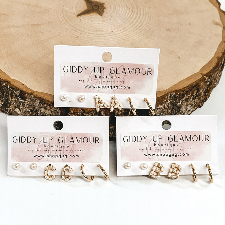 This picture includes three earring initial sets. Each set includes pearl studs, pearl initial studs, and gold hoop huggies. There is one set on a piece of wood and the other two are in front of the piece on wood. All is pictured on a white background.