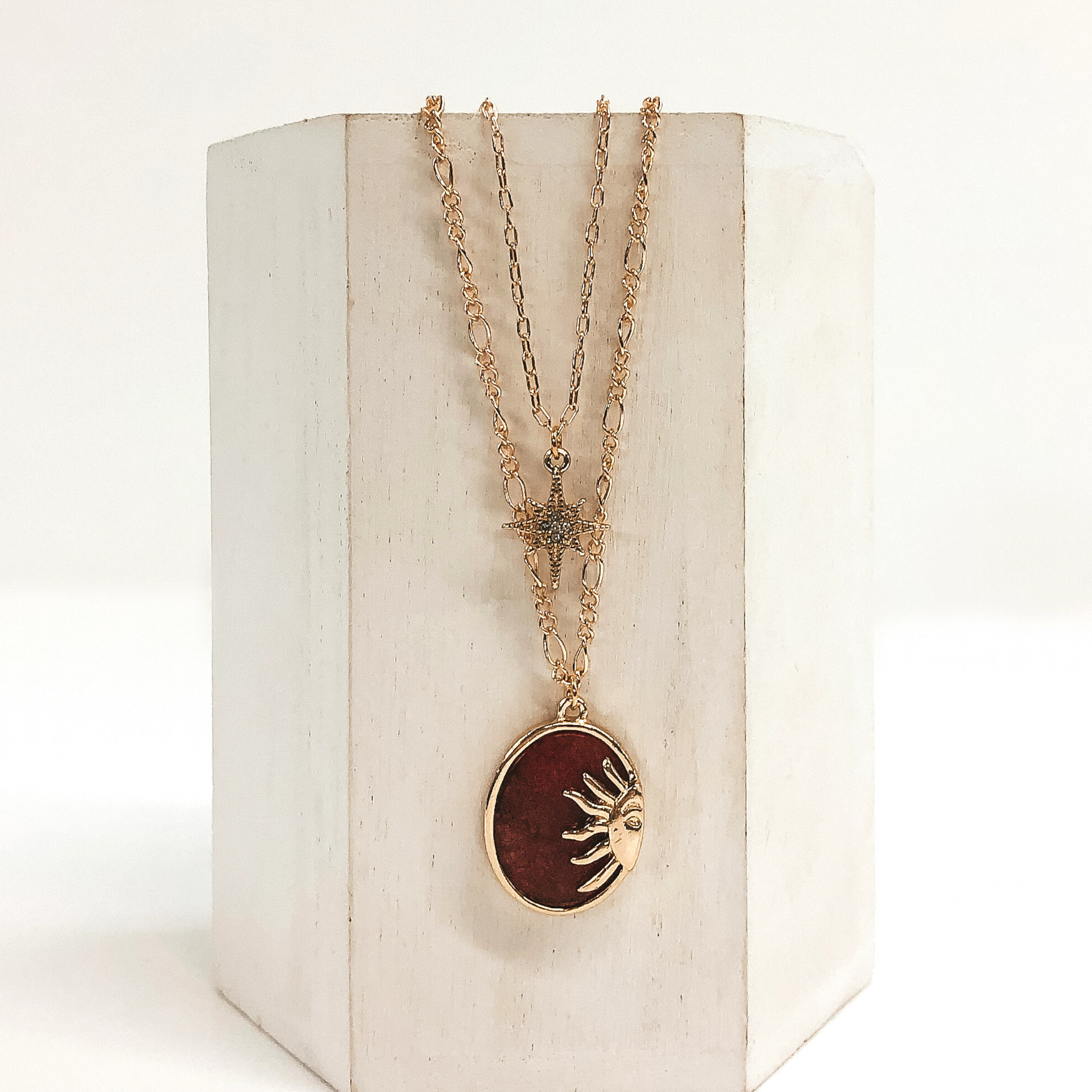 Double layered chain necklace in gold. The shorter strand has a gold star pendant with a center clear crystal and the longer strand has an oval burgundy stone pendant with a half sun gold charm. This necklace is pictured hanging on a white block on a white background. 