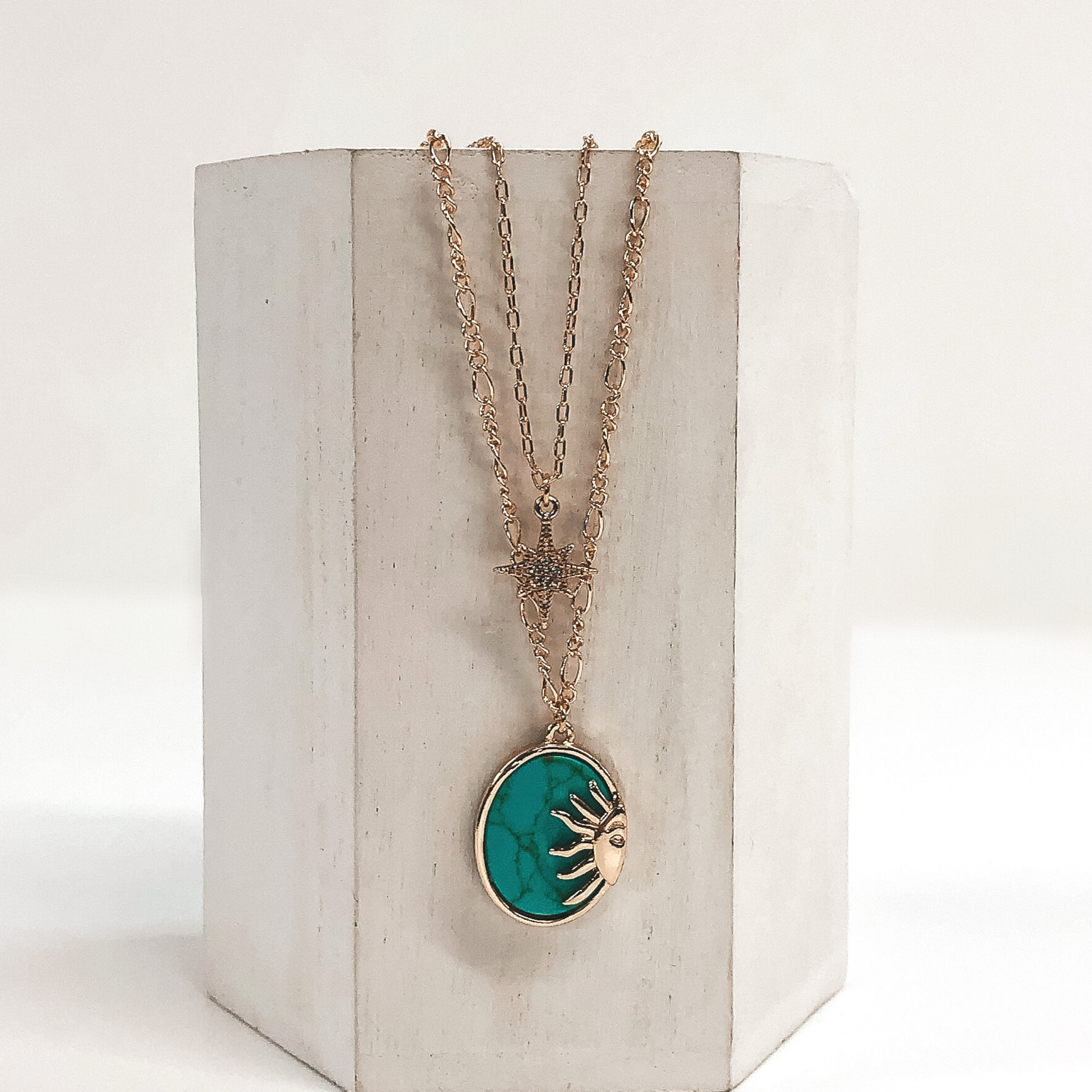 Double layered chain necklace in gold. The shorter strand has a gold star pendant with a center clear crystal and the longer strand has an oval turquoise stone pendant with a half sun gold charm. This necklace is pictured hanging on a white block on a white background.