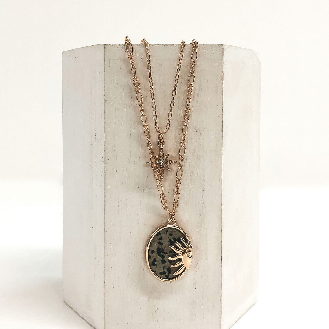 Double layered chain necklace in gold. The shorter strand has a gold star pendant with a center clear crystal and the longer strand has an oval dotted stone pendant with a half sun gold charm. This necklace is pictured hanging on a white block on a white background. 