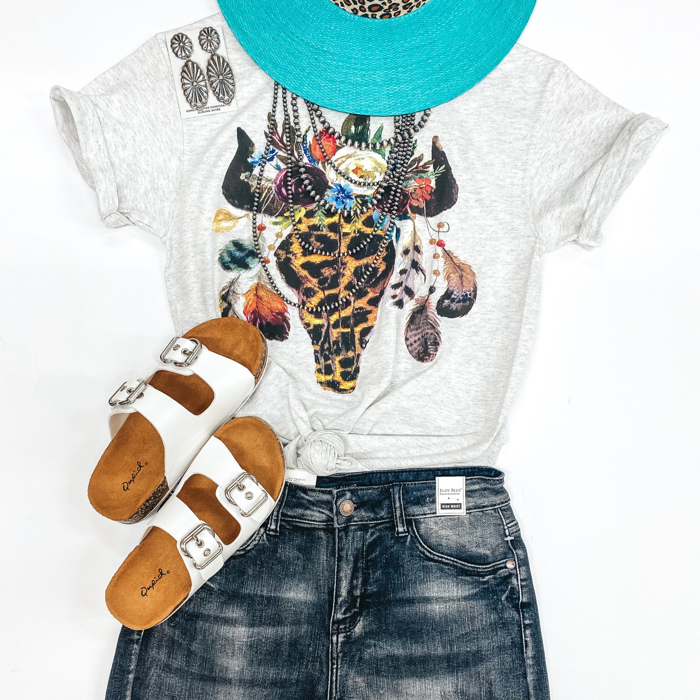 A heather grey tee shirt with a leopard print bull skull with flowers and feathers. Pictured with a turquoise straw hat, genuine Navajo jewelry, white sandals, and denim shorts.