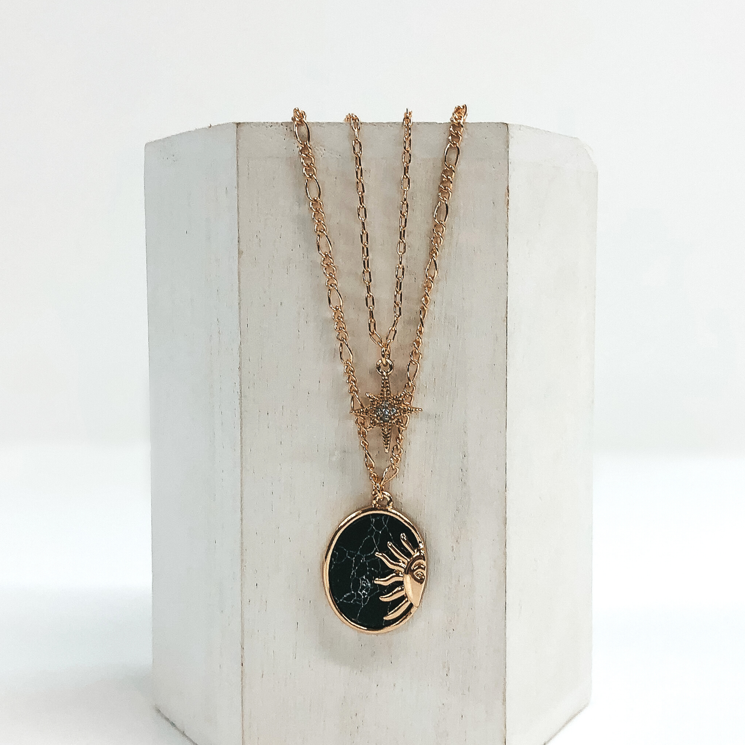 Double layered chain necklace in gold. The shorter strand has a gold star pendant with a center clear crystal and the longer strand has an oval black stone pendant with a half sun gold charm. This necklace is pictured hanging on a white block on a white background. 