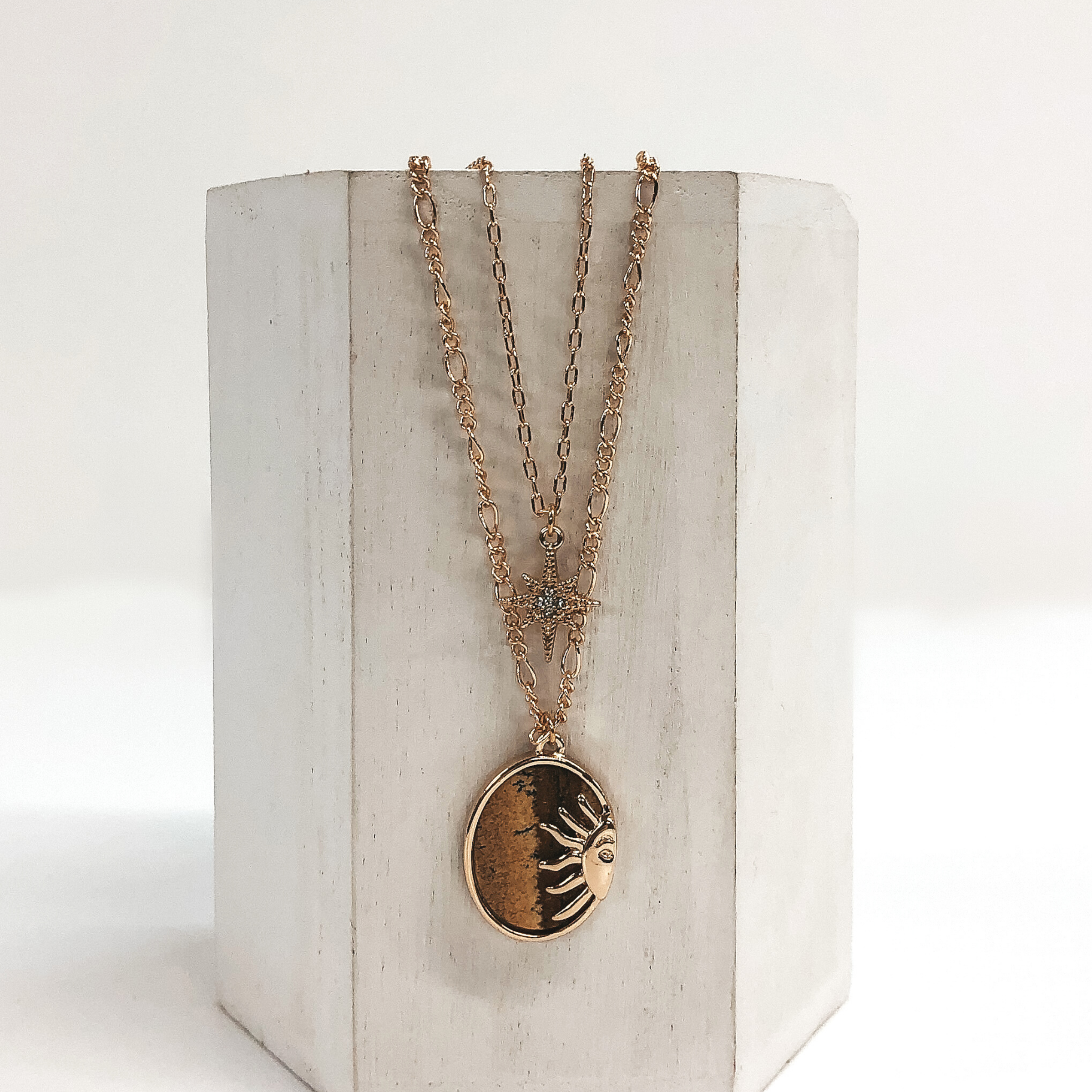 Double layered chain necklace in gold. The shorter strand has a gold star pendant with a center clear crystal and the longer strand has an oval tan stone pendant with a half sun gold charm. This necklace is pictured hanging on a white block on a white background. 