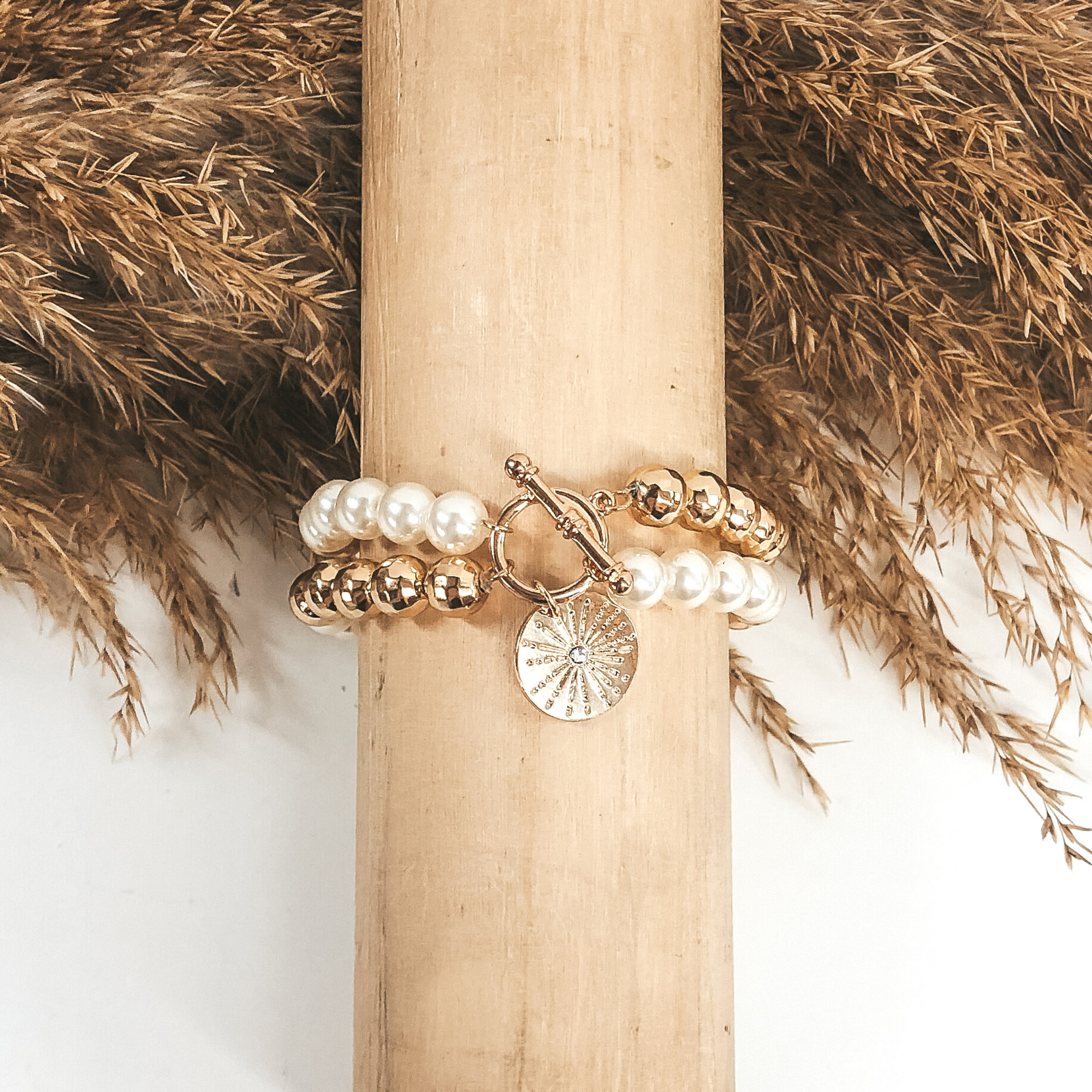 This is a two stranded bracelet that includes segments of gold or white beads connect with a toggle clasp. There is a gold circle pendant with a little sun design and a tiny center crystal. This bracelet is pictured wrapped on a wooden bracelet holder on a white background with tan floral in the top half of the picture.