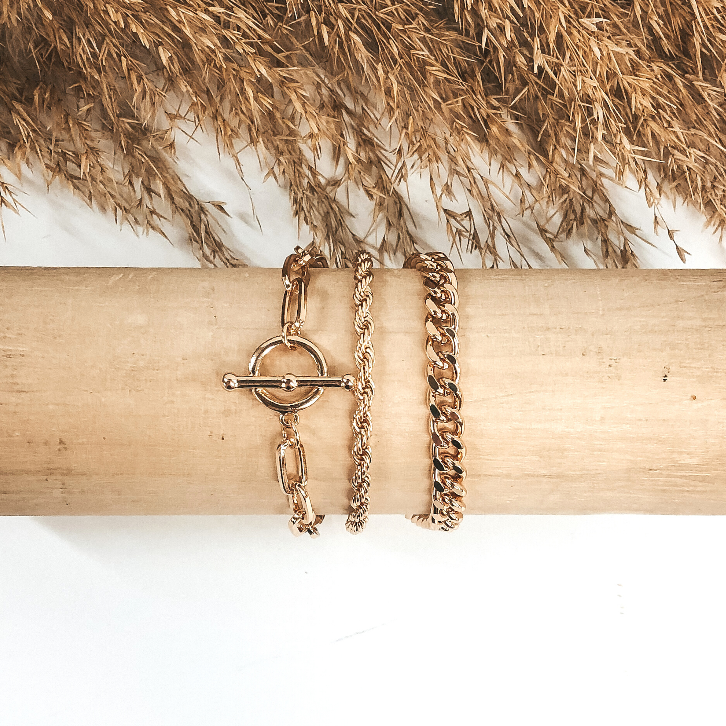 This is a three stranded gold, magnetic bracelet that includes a curb chain, a rope chain, and a paperclip chain with a toggle clasp pendant. This bracelet is pictured wrapped around a wooden bracelet holder on a white background with tan floral at the top of the picture. 