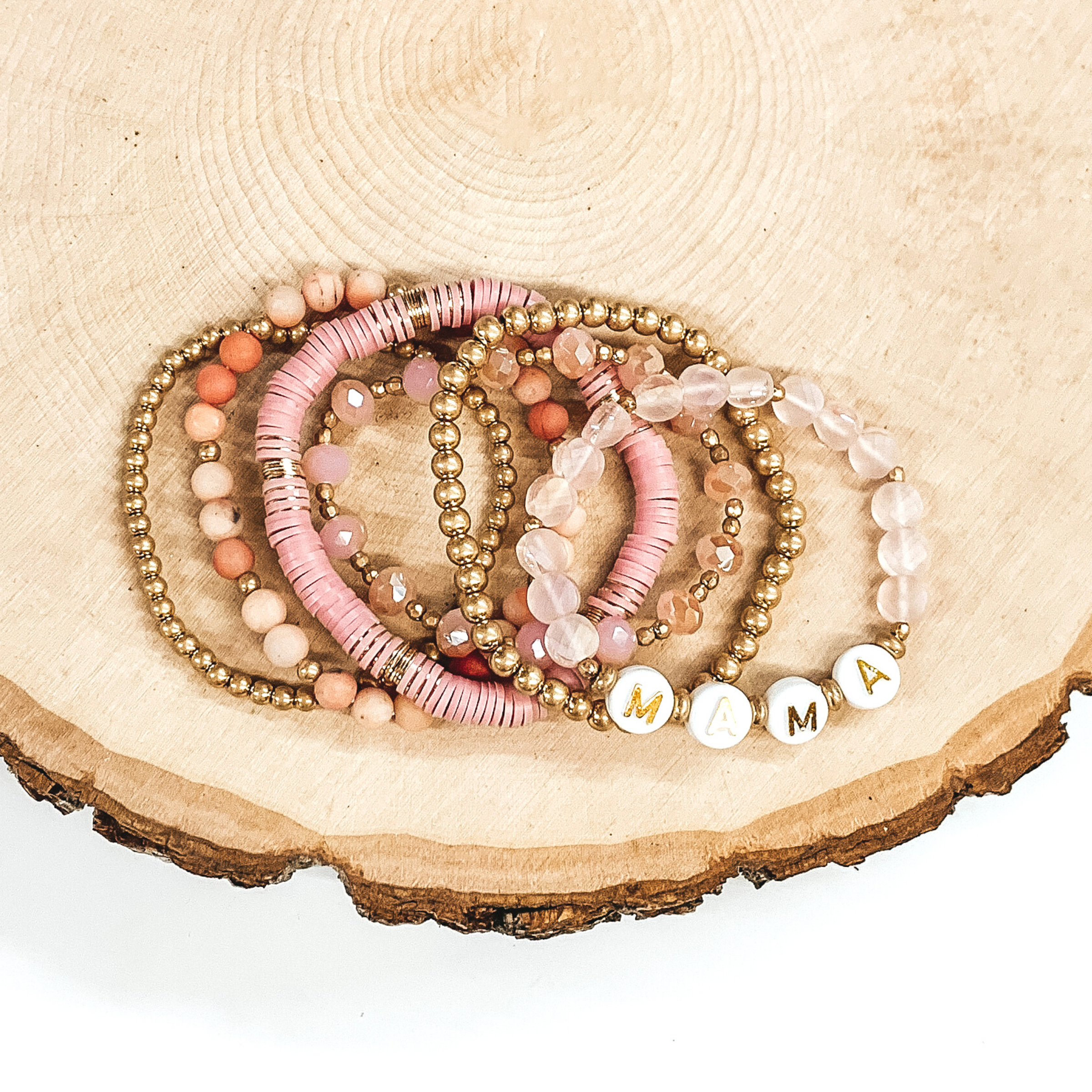 This bracelet set has a mix of light pink disk beaded bracelet, light pink circle beaded bracelet, two gold beaded bracelets, a light pink crystal beaded bracelet. One of the crystal beaded bracelets has letter beads that spell out "MAMA." These bracelets are pictured on a piece of wood on a white background.