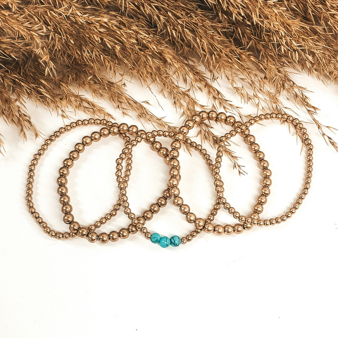 This is a gold beaded bracelet set that has different sizes of beads. One bracelet includes three turquoise beads that are lined up together to add a pop of color. These bracelets are laid on top of each other to be pictured on a white background with tan floral at the top of the picture. 