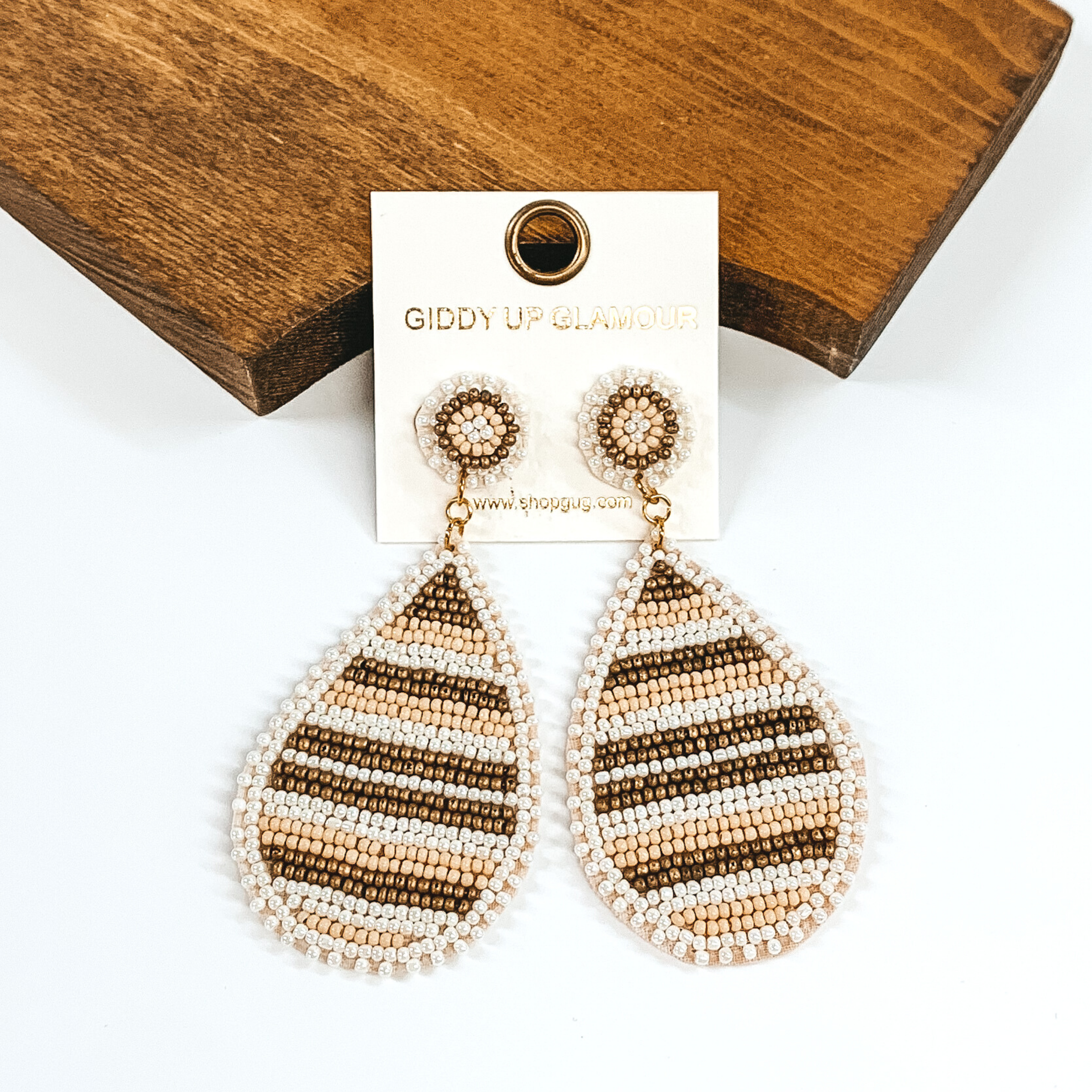 These earrings have a circle beaded stud that has a beaded teardrop hanging part. This pair of earrings is striped and includes white, beige, and gold colors. These earrings are pictured on a white background and laying against a dark piece of wood. 