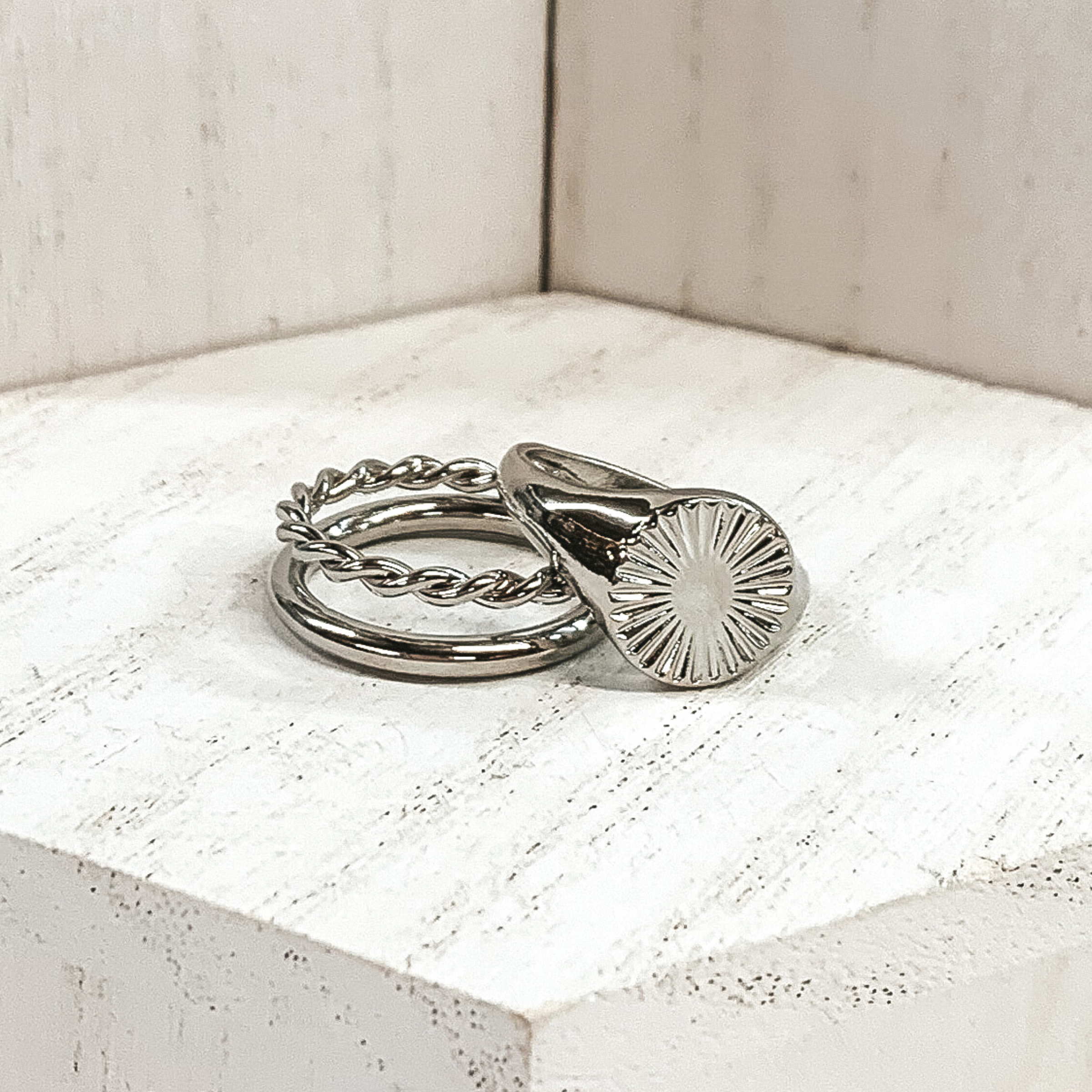 Both rings are silver in color.  The double ring has one plain band and one twisted band. The ring that is leaning on it has a circle pendant with a starburst design on it. These rings are pictured on a white background.