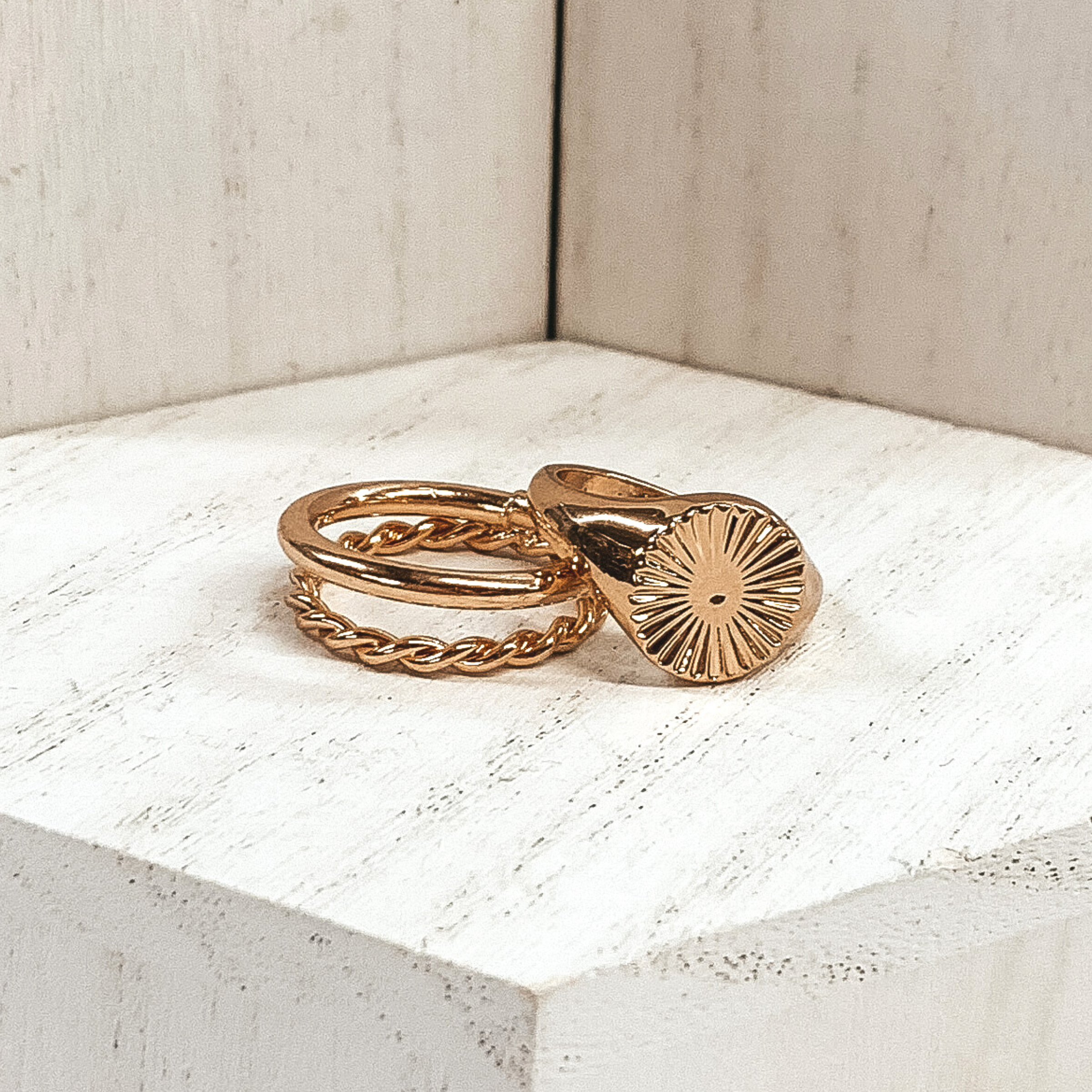 Both rings are gold in color. The double ring has one plain band and one twisted band. The ring that is leaning on it has a circle pendant with a starburst design on it. These rings are pictured on a white background.