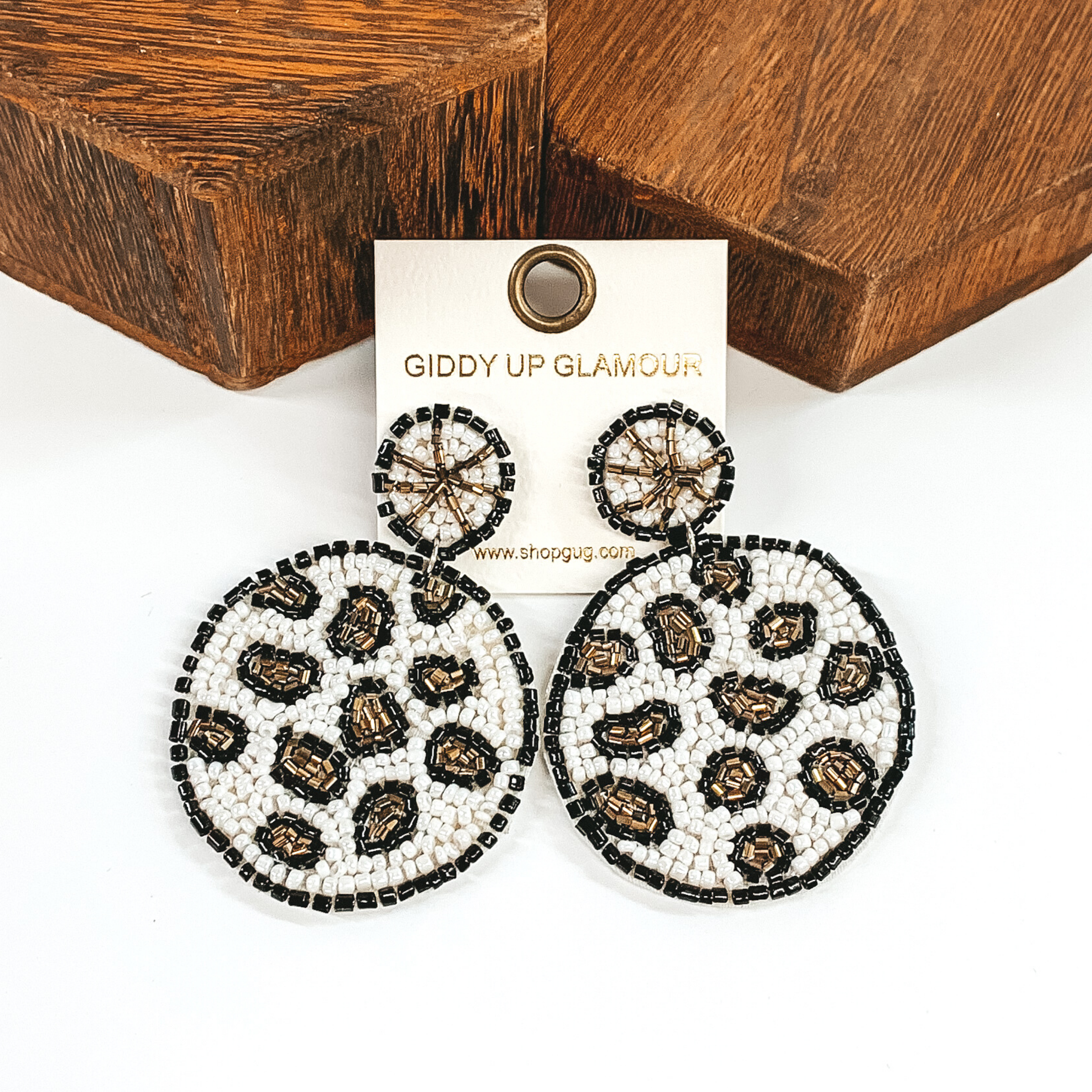 The studs are a small beaded circle with a hanging larger beaded circle. These beaded earrings includes the colors white, black, and gold in a leopard design. These earrings are pictured on a white background with brown blocks behind the earrings. 