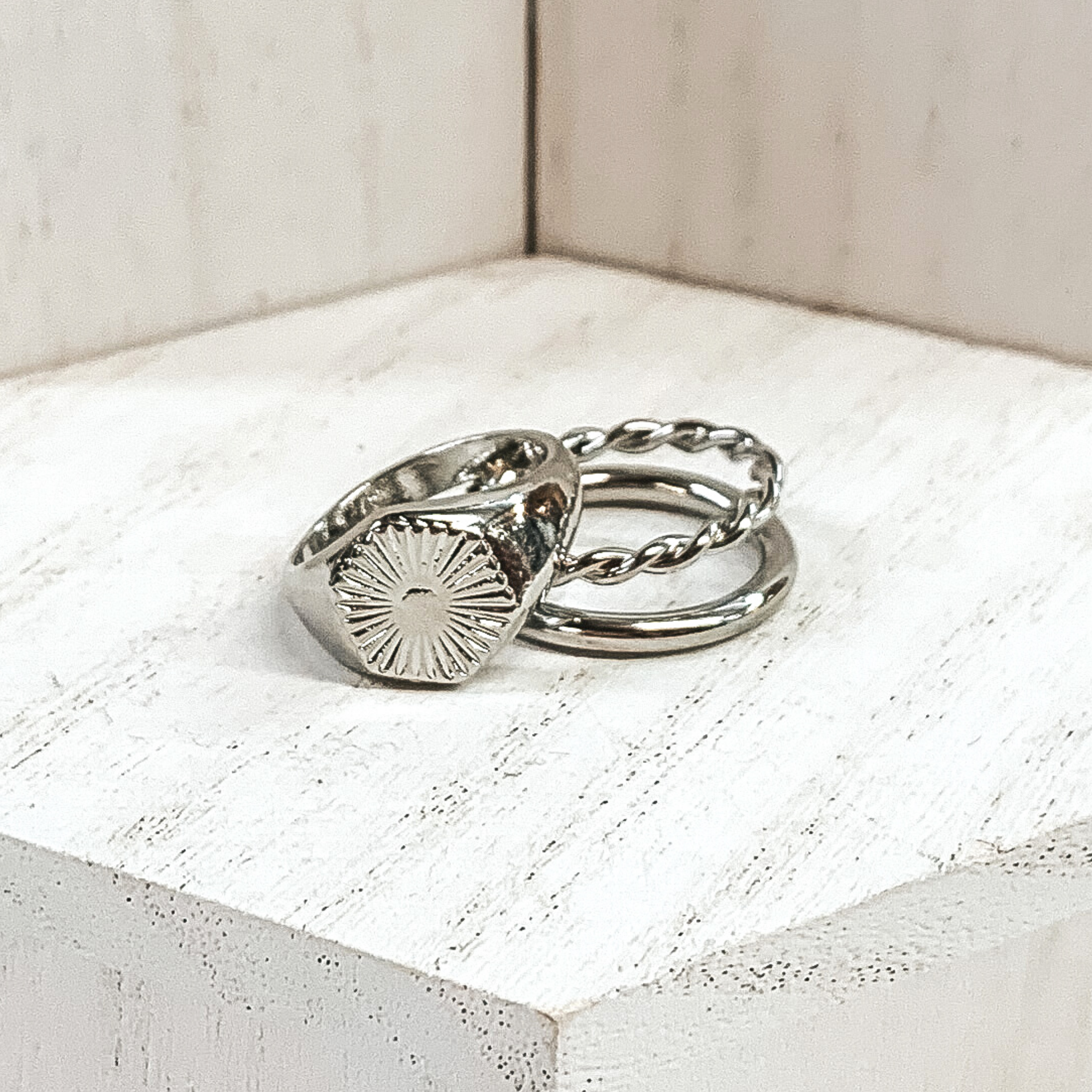 Both rings are silver in color. The double ring has one plain band and one twisted band. The ring that is leaning on it has a hexagon pendant with a starburst design on it. These rings are pictured on a white background.