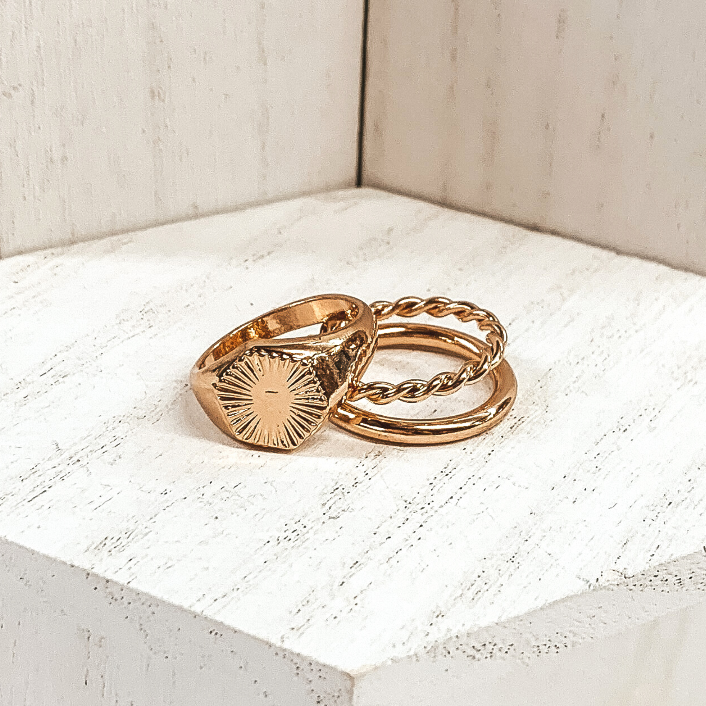 Both rings are gold in color. The double ring has one plain band and one twisted band. The ring that is leaning on it has a hexagon pendant with a starburst design on it. These rings are pictured on a white background.